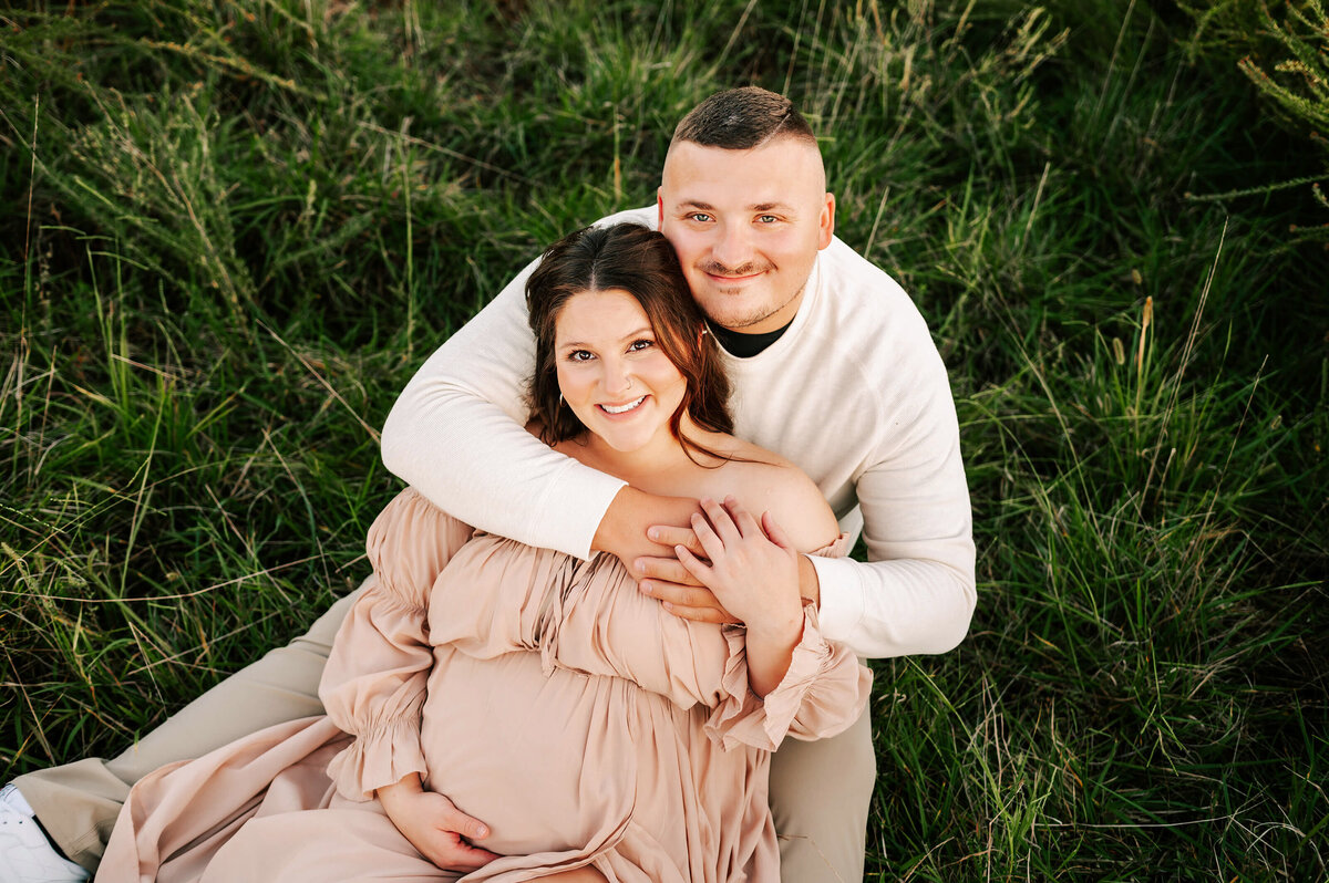 pregnant couple smiling sitting in grass captured by Joplin MO maternity photographer The XO Photography