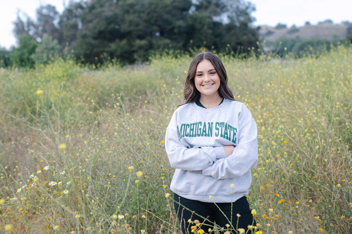A person stands in a field of wildflowers, smiling with arms crossed. They are wearing a gray Michigan State sweatshirt. Trees and hills are visible in the background under a cloudy sky.