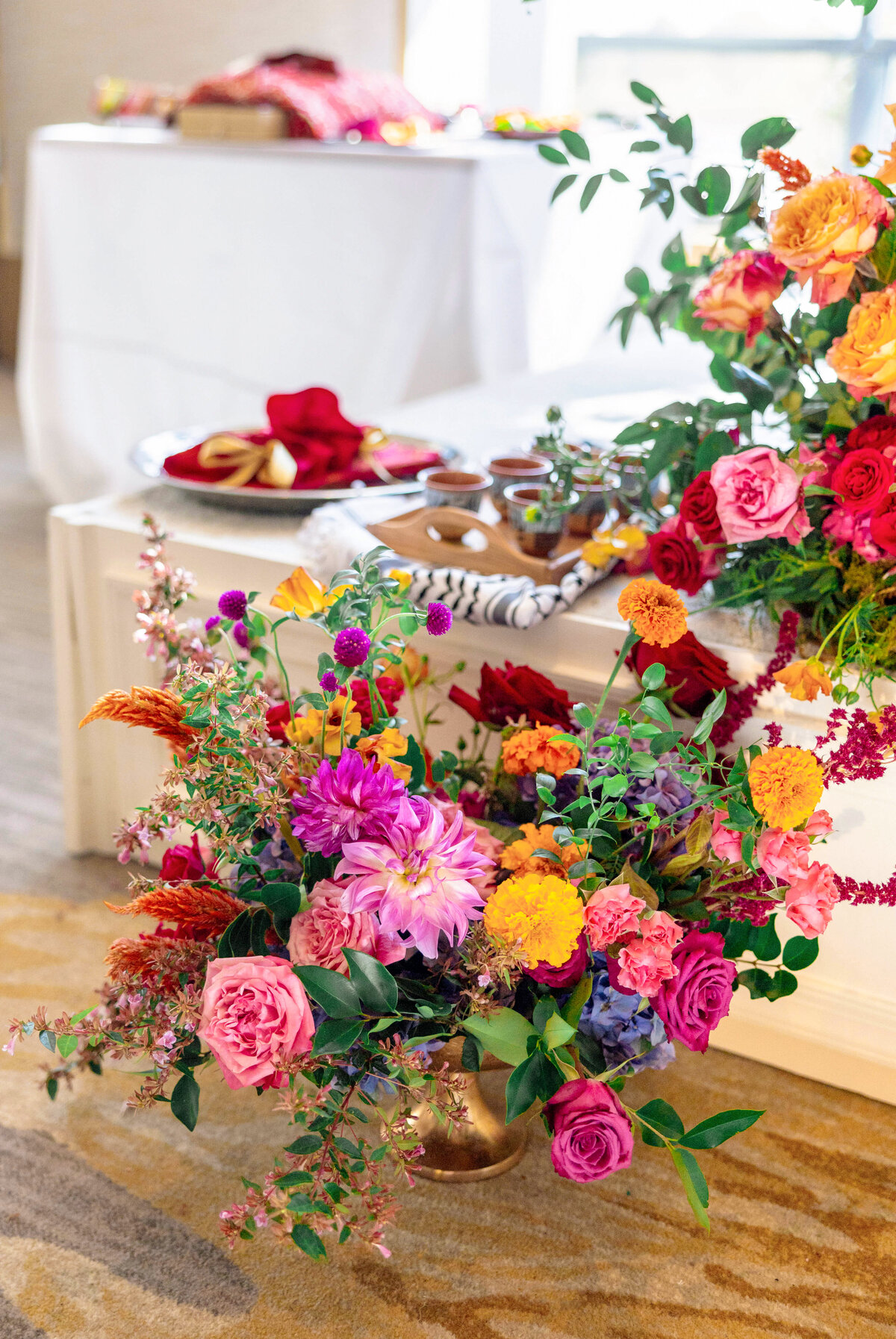 A vibrant floral arrangement with pink, purple, orange, and red flowers placed in front of a table set with plates and food. The scene suggests a festive or decorative indoor setting.