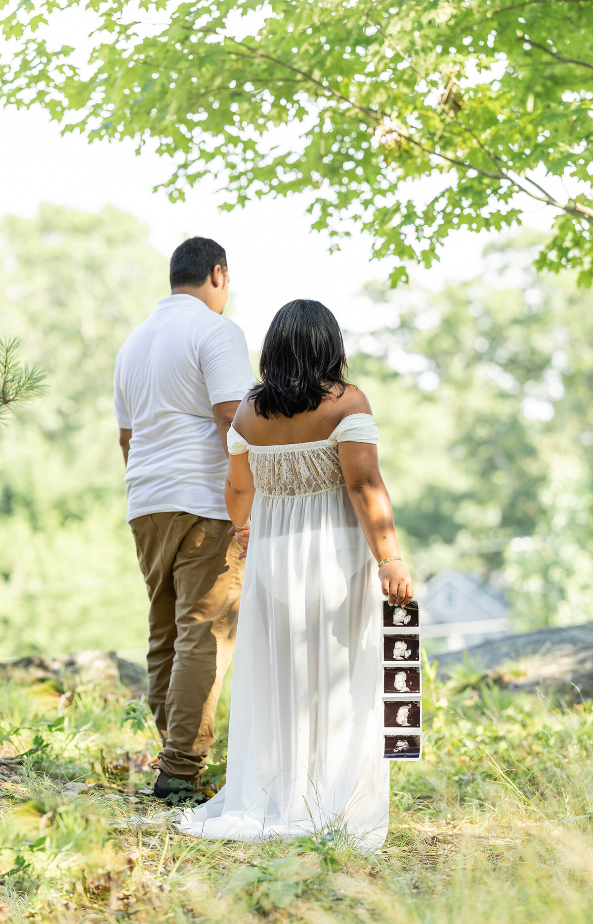 Expecting parents stand towards the cliff with their backs to the camera  while holding their sonogram during their maternity session in Connecticut