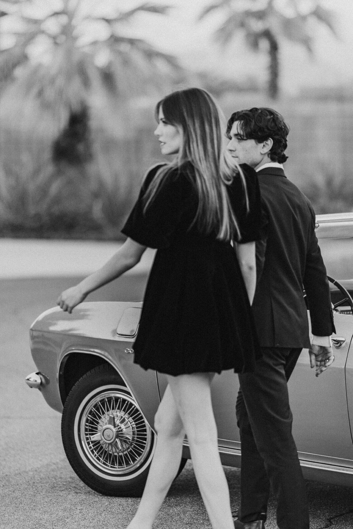Black and white photo of couple walking side by side near vintage car, showing movement, connection, and quiet energy.