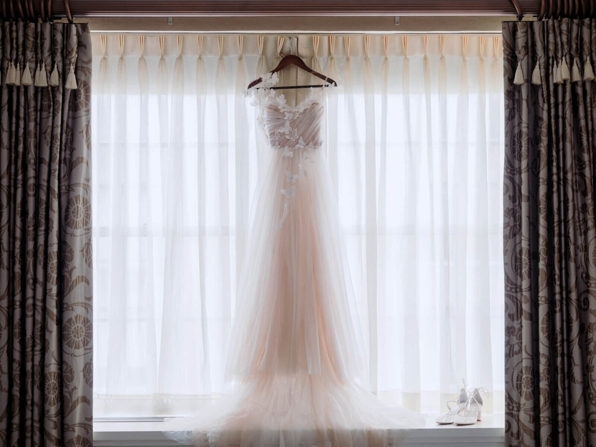 A wedding dress hanging in the middle of a window with a white and brown curtain and wedding shoes at the bottom