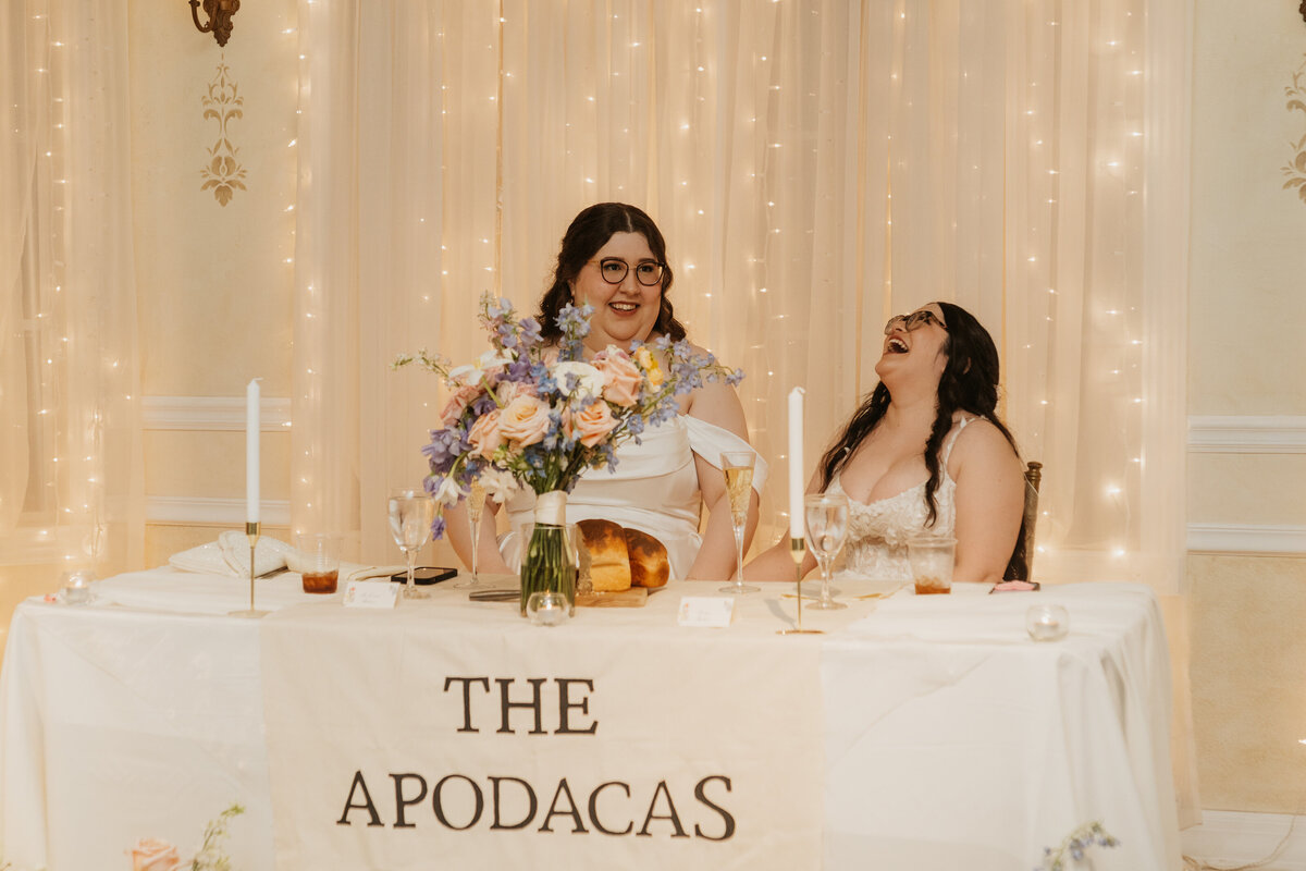 A newlywed couple laughing at sitting at a table at their reception 