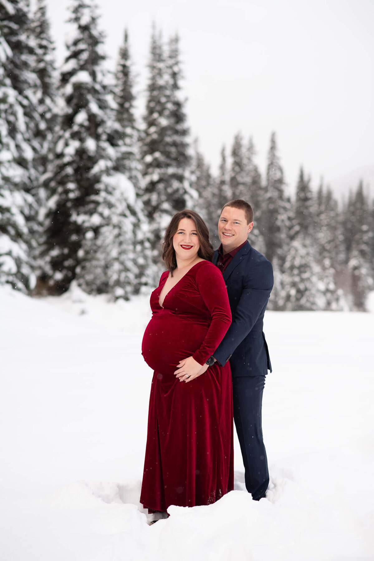 A husband and wife are in and embrace with a snowy background. She wears a red dress and he wears a suit. Captured by Brittany Danielle
