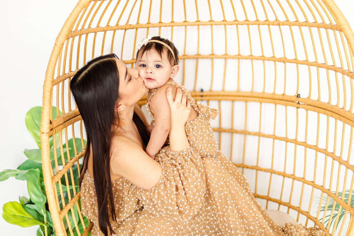 A mother sitting in a boho egg chair, lifting her infant for a kiss while the baby gazes directly into the camera with wide, curious eyes.