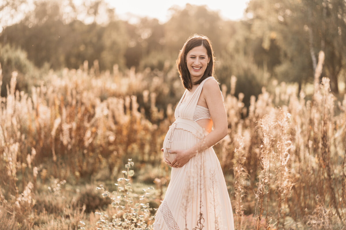 Photo of a pregnant woman standing in a long field of grass and flowers at sunset