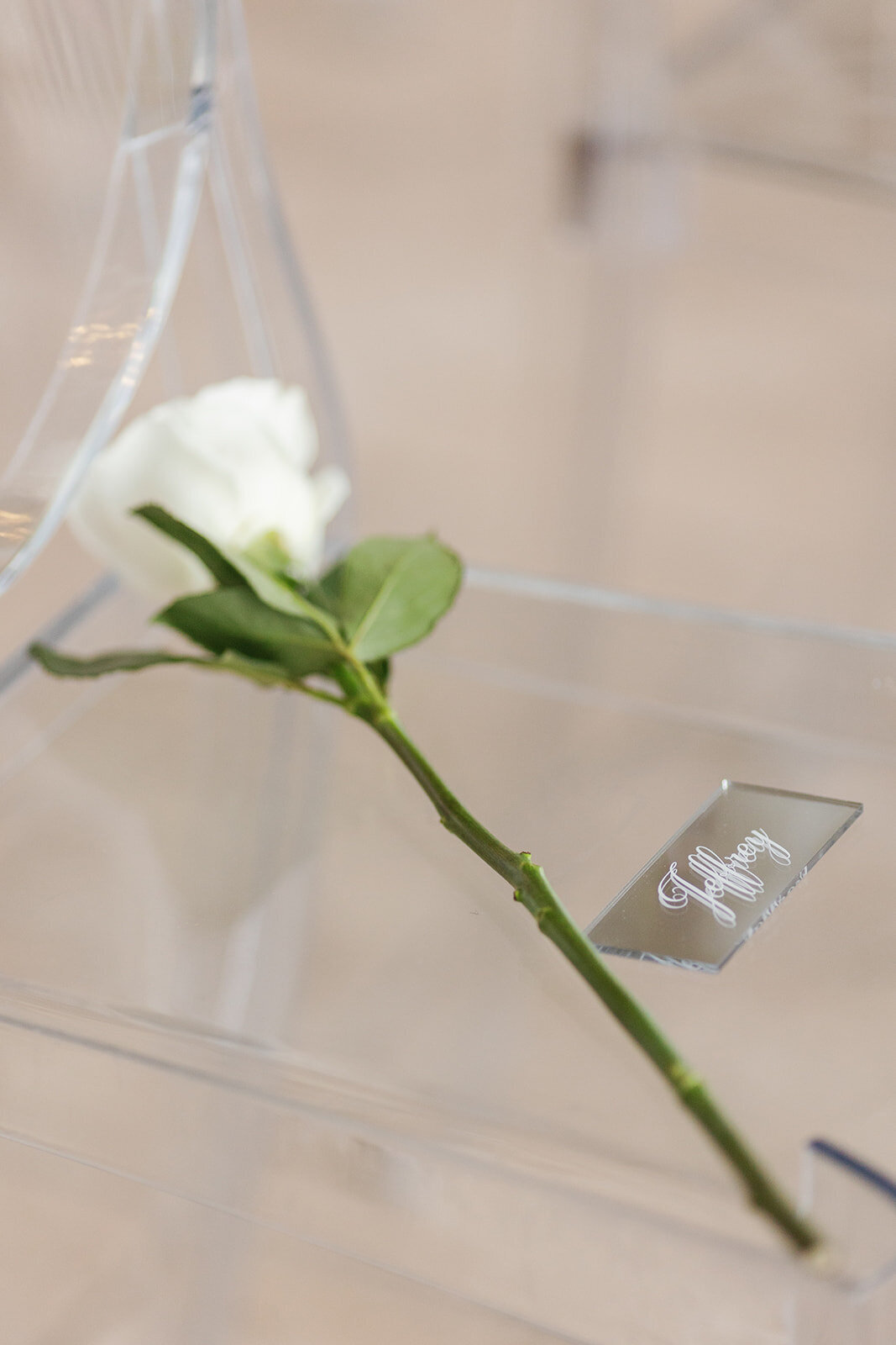 A single white rose and acrylic name tag delicately placed on a clear wedding chair during an Ocean Reef wedding in Key Largo, captured by Claudia Amalia Photography.