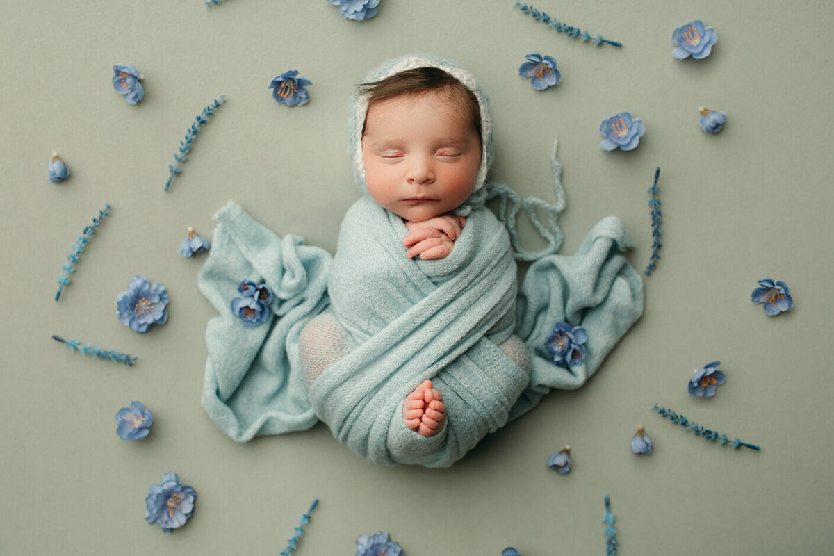 baby girl wearing blue asleep with toes peeking out of a blue wrap surrounded by blue flowers and a blue bonnet