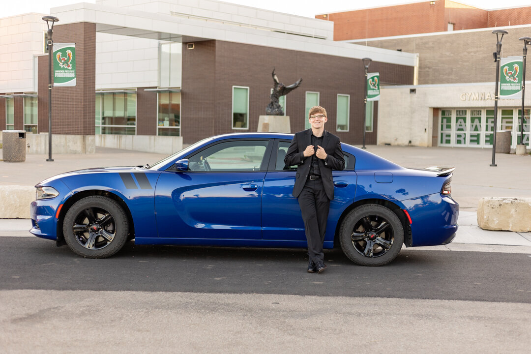 A senior guy standing leaning against his car grabbing his suite jacket in front of his high school in Lawrence, KS