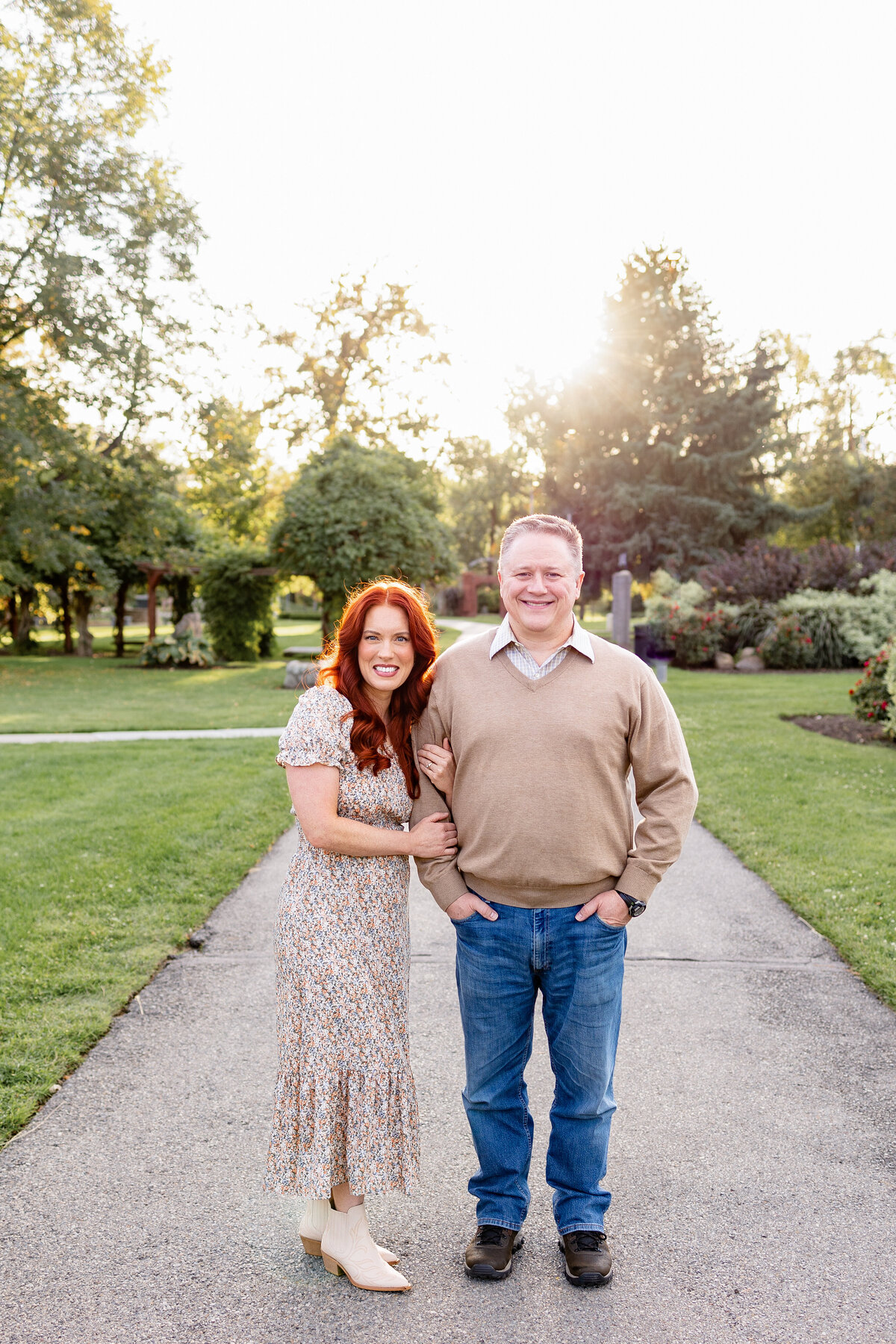A couple poses together on a paved path in a lush green park, with the sun setting behind them. The woman, with red hair, leans on her partner’s arm while both smile warmly at the camera.