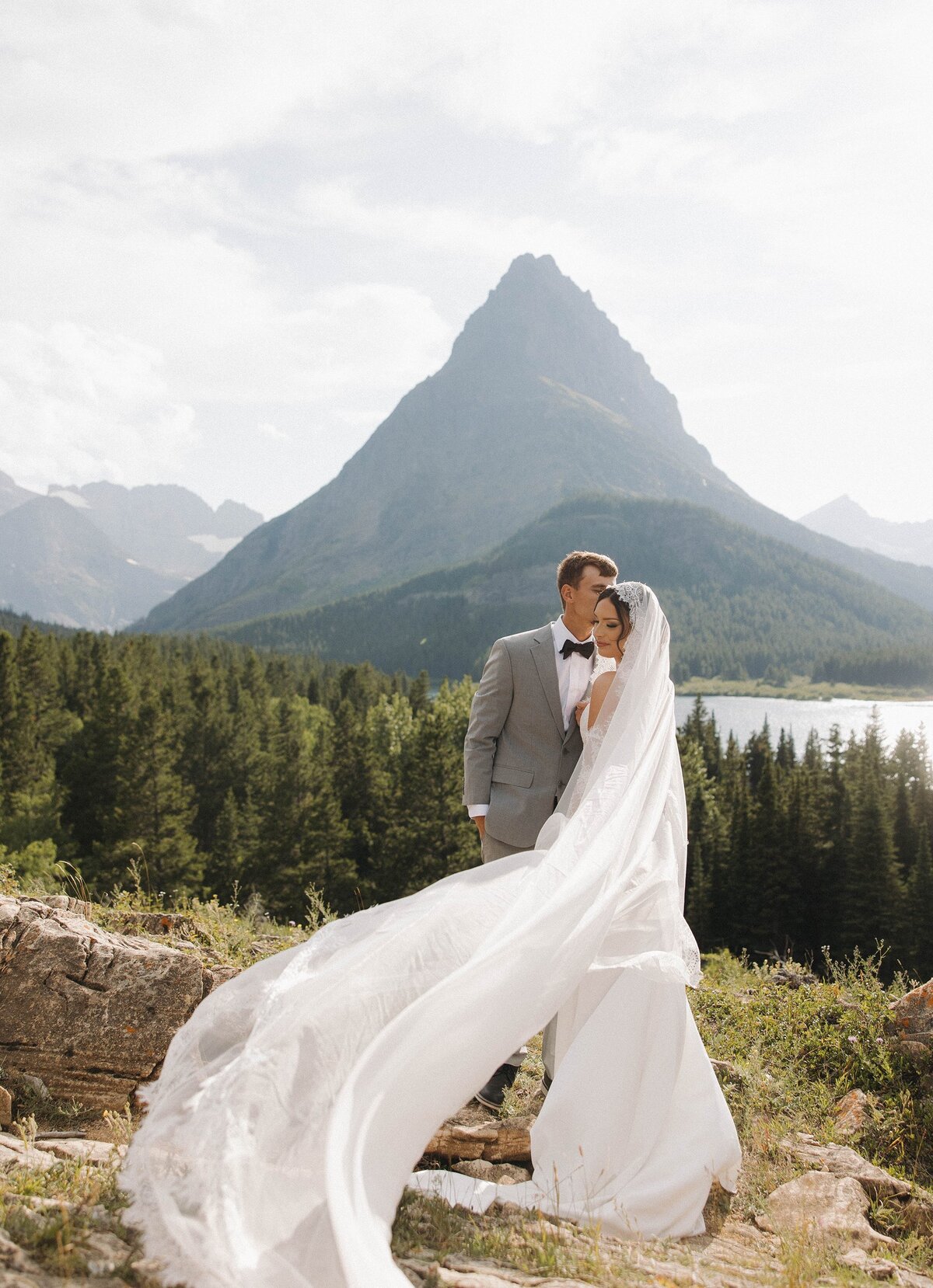Bride and groom embracing outdoors with mountain backdrop and flowing veil.