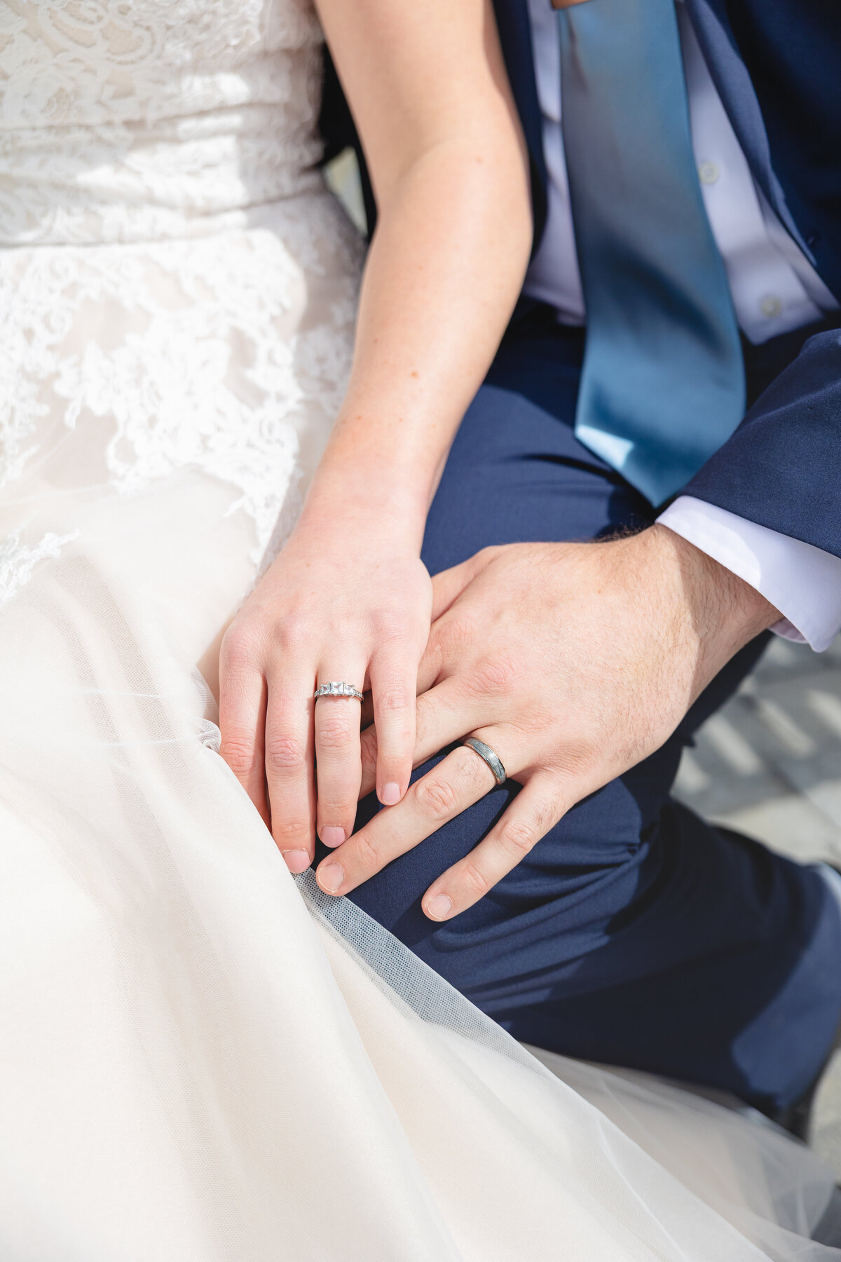 Close-up of bride and groom’s hands resting together, showing their wedding rings against the backdrop of the bride’s lace gown and groom’s navy blue suit.