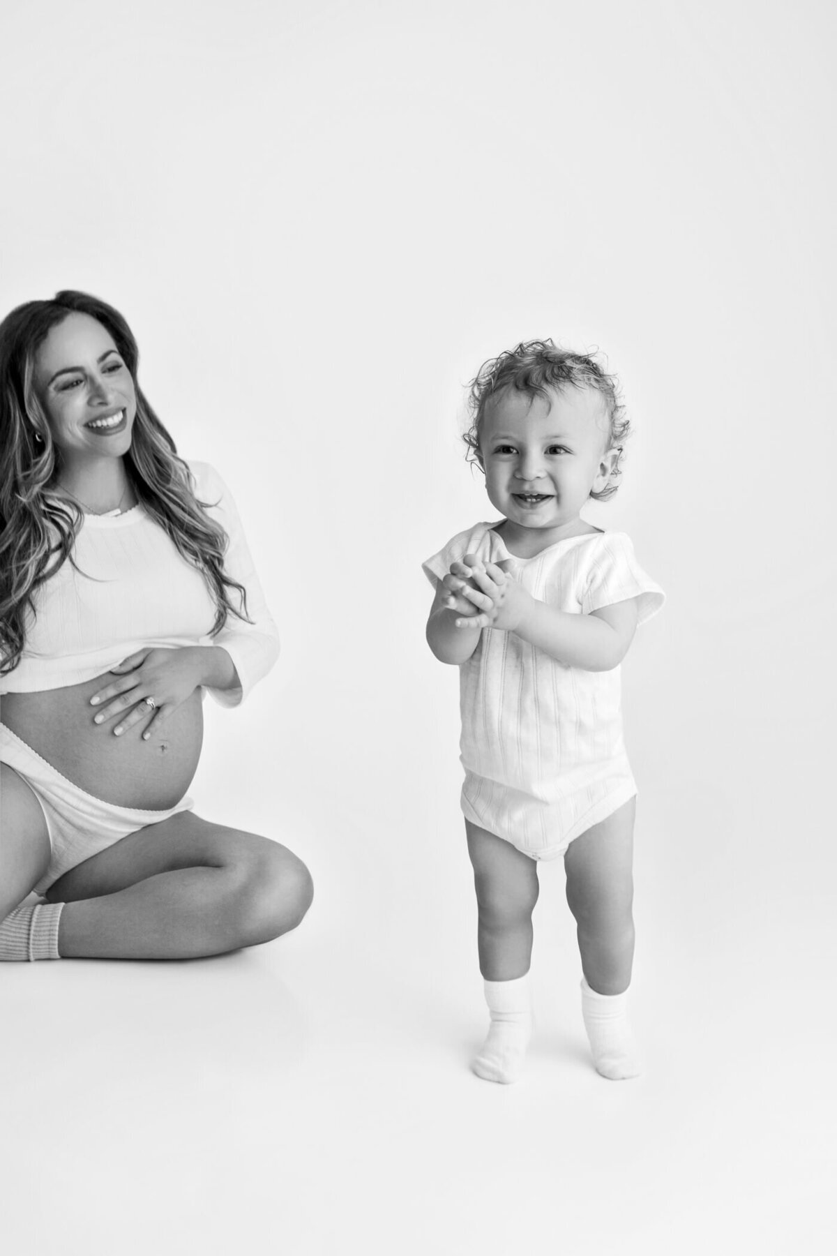 A joyful black-and-white maternity portrait featuring a pregnant woman sitting on the floor, smiling as she watches her toddler stand and clap their hands. The woman wears a white long-sleeve top and underwear, gently cradling her baby bump with one hand. The toddler, dressed in a white onesie and socks, stands confidently in front of her, exuding happiness and energy. The minimalistic background enhances the focus on the loving interaction between mother and child.