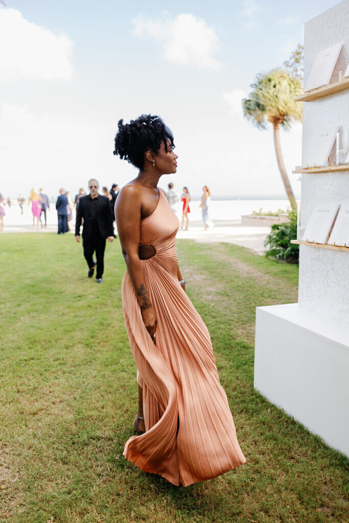 A stylish wedding guest in a flowing rust-colored gown checks the seating chart during an elegant Florida Keys wedding. Captured by Claudia Amalia Photography.