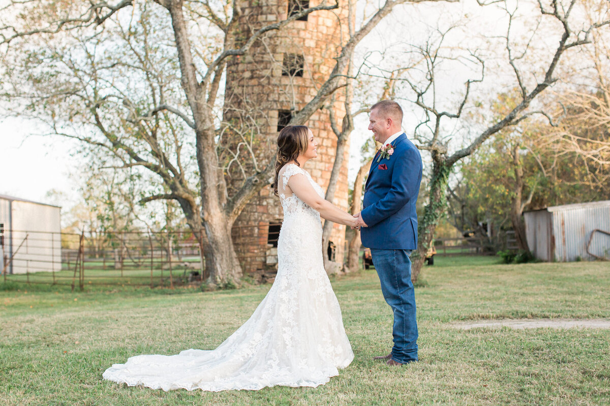 The newly married couple stand facing each other, holding hands and smiling broadly in front of an old grain silo