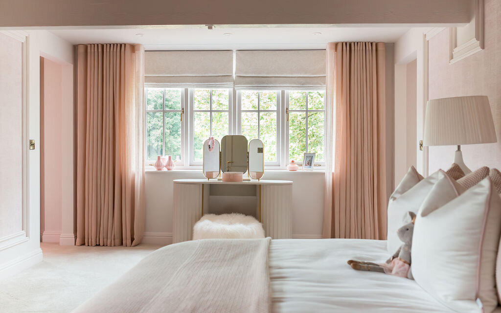 A cozy bedroom with soft pastel decor. The bed is dressed in white and pink linens and fluffy pillows. At the end of the room, a window with pink curtains brings in natural light, and beneath it is a vanity table with a mirror and decorative items.