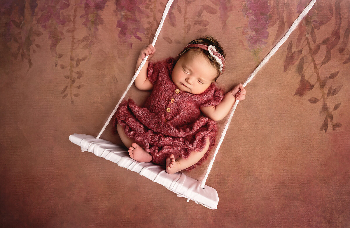 Infant girl in a pink knit dress sitting on a white swing in studio.