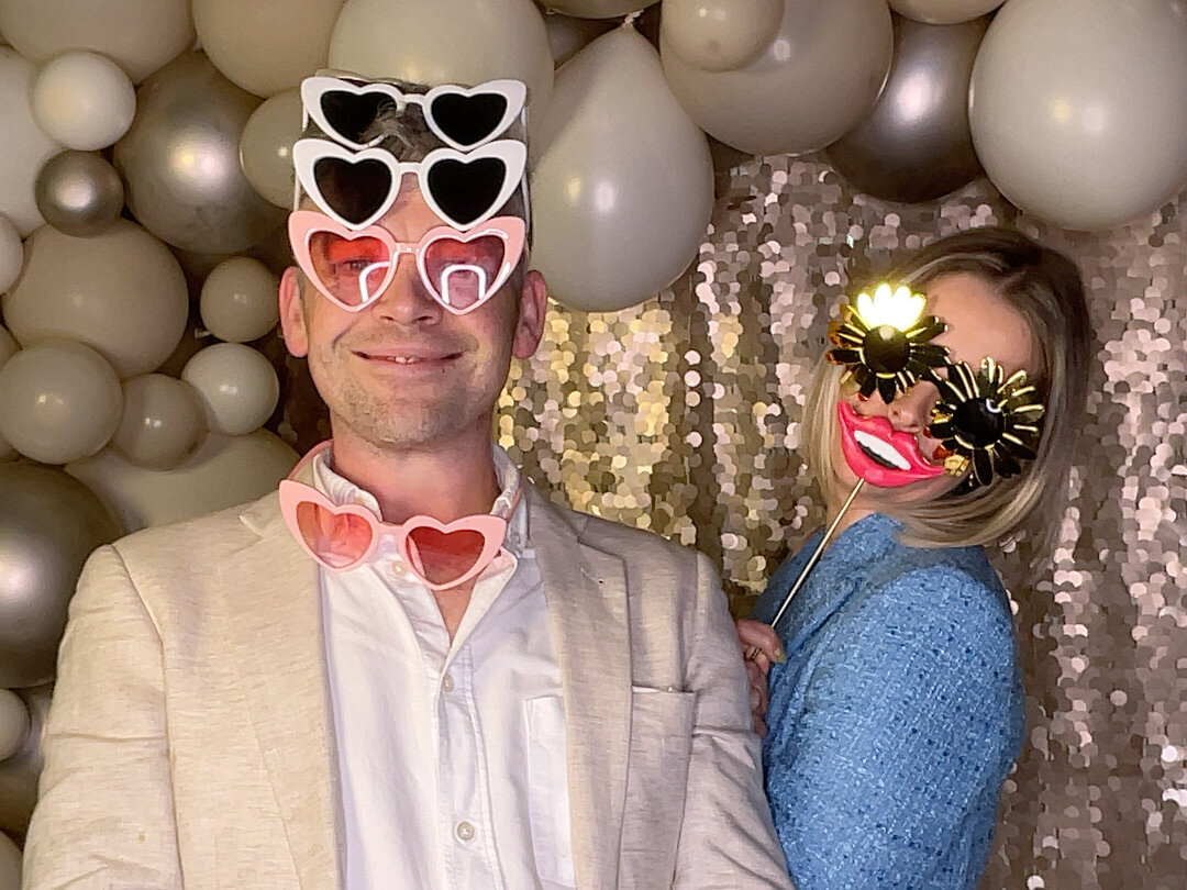 two young people with heart sunglasses in front of a gold and champagne sequin backdrop in a photo booth