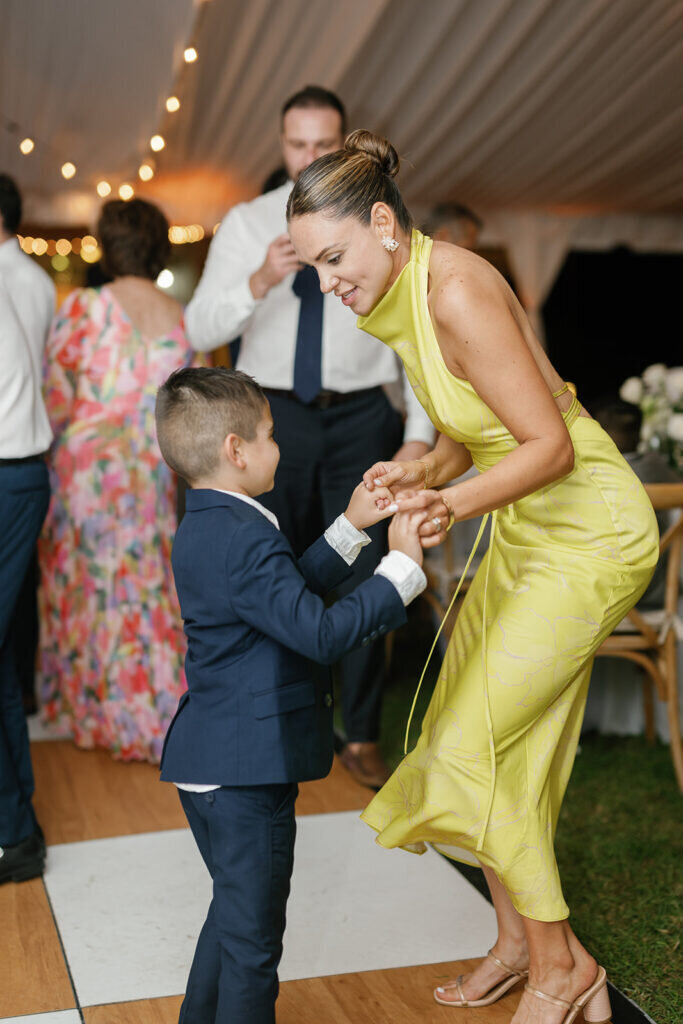 A heartwarming wedding reception moment as a stylish guest in a yellow dress dances with a young boy in a navy suit, sharing a joyful connection under twinkling lights. Captured by Claudia Amalia Photography, a Florida Keys wedding photographer specializing in destination wedding photography.
