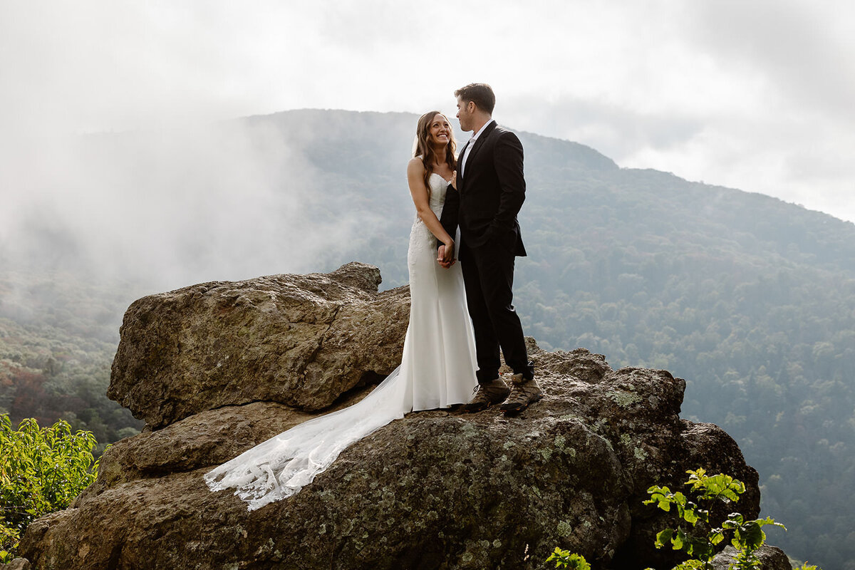A newlywed couple holding hands and standing on a small boulder together