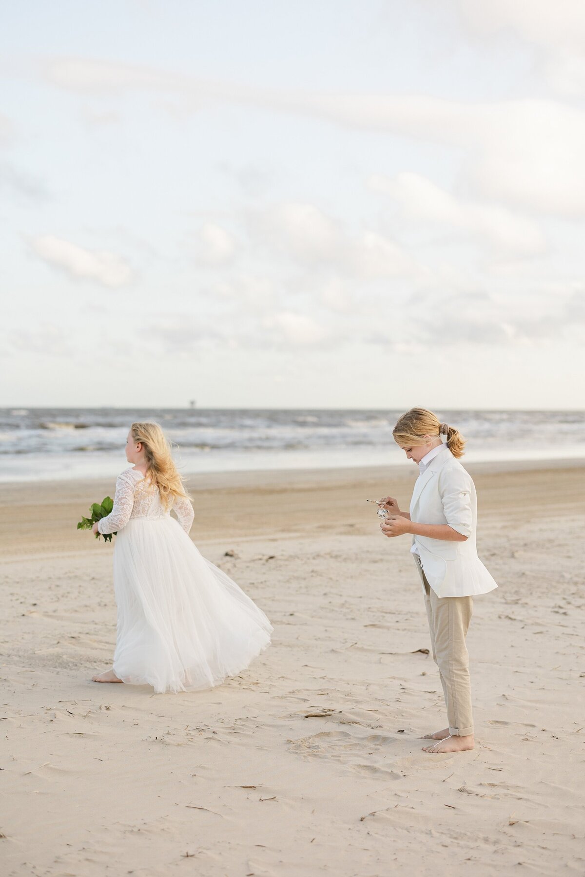 Candid shot of two child attendants waiting patiently on the beach before a wedding ceremony at Crystal Beach, Texas. The child on the left is wearing a flowing, white dress, is holding a bouquet, and is looking out at the ocean. The child on the right is wearing a jacket, khaki pants, and is looking down at something in their hands.