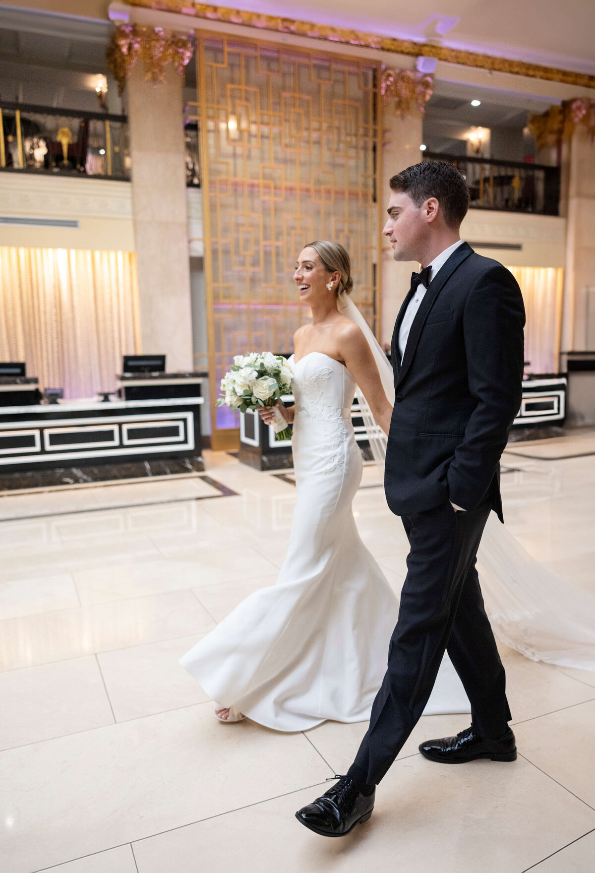 A bride in a strapless white gown holds a bouquet while walking with a groom in a black tuxedo. They are in an elegant venue with marble floors and ornate golden decor in the background.