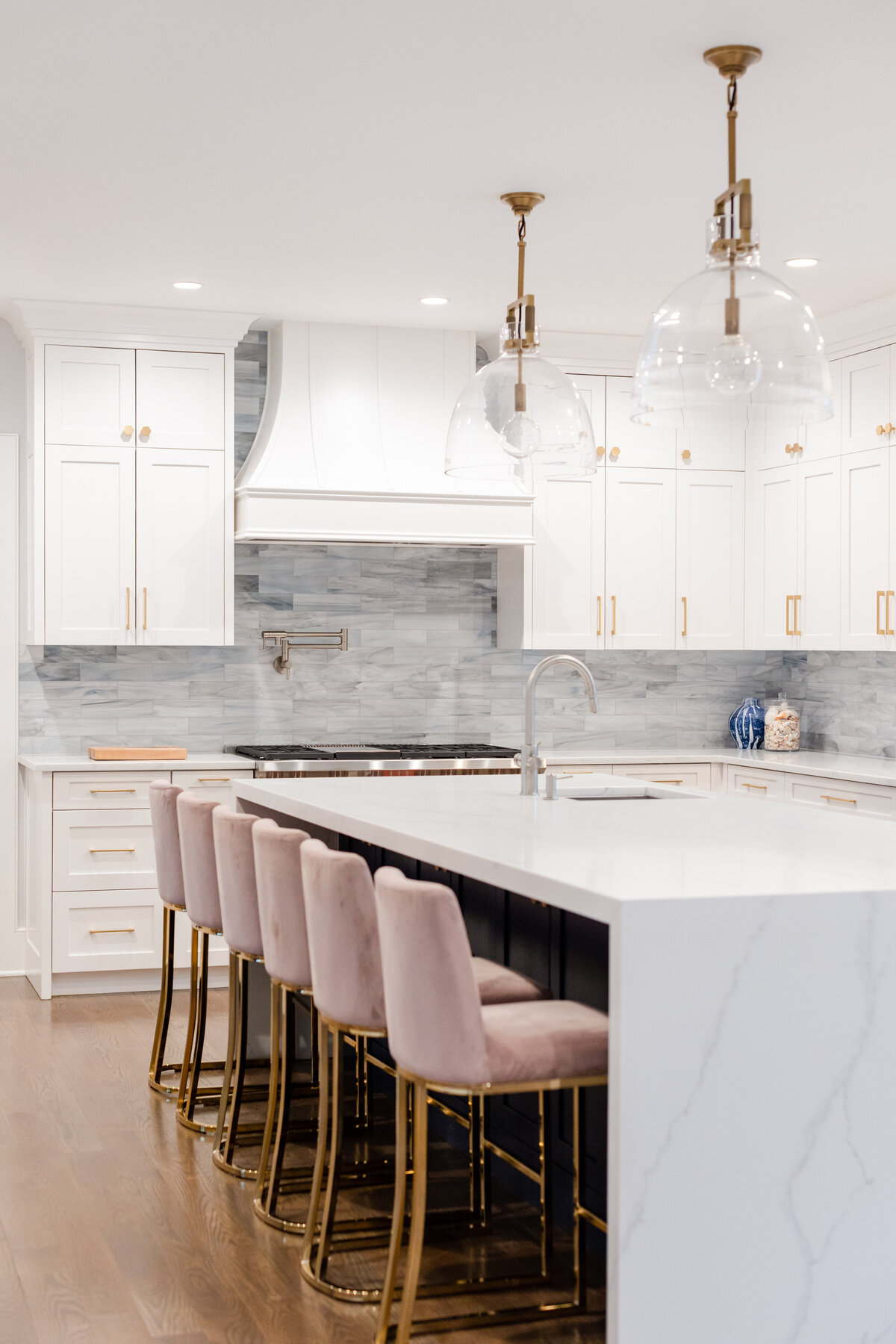 The kitchen includes white cabinetry, a grey backsplash, gold hardware, and a waterfall island with pink barstools. The Mingus blue backsplash is hand-cut glass tile, and the island features a waterfall-edge quartz countertop.