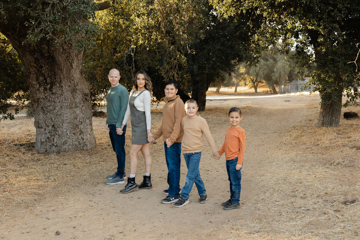 Family of four holding hands and smiling in front of large tree. Photographed by Yucaipa family photographer Kaitlyn Dawn Photography.