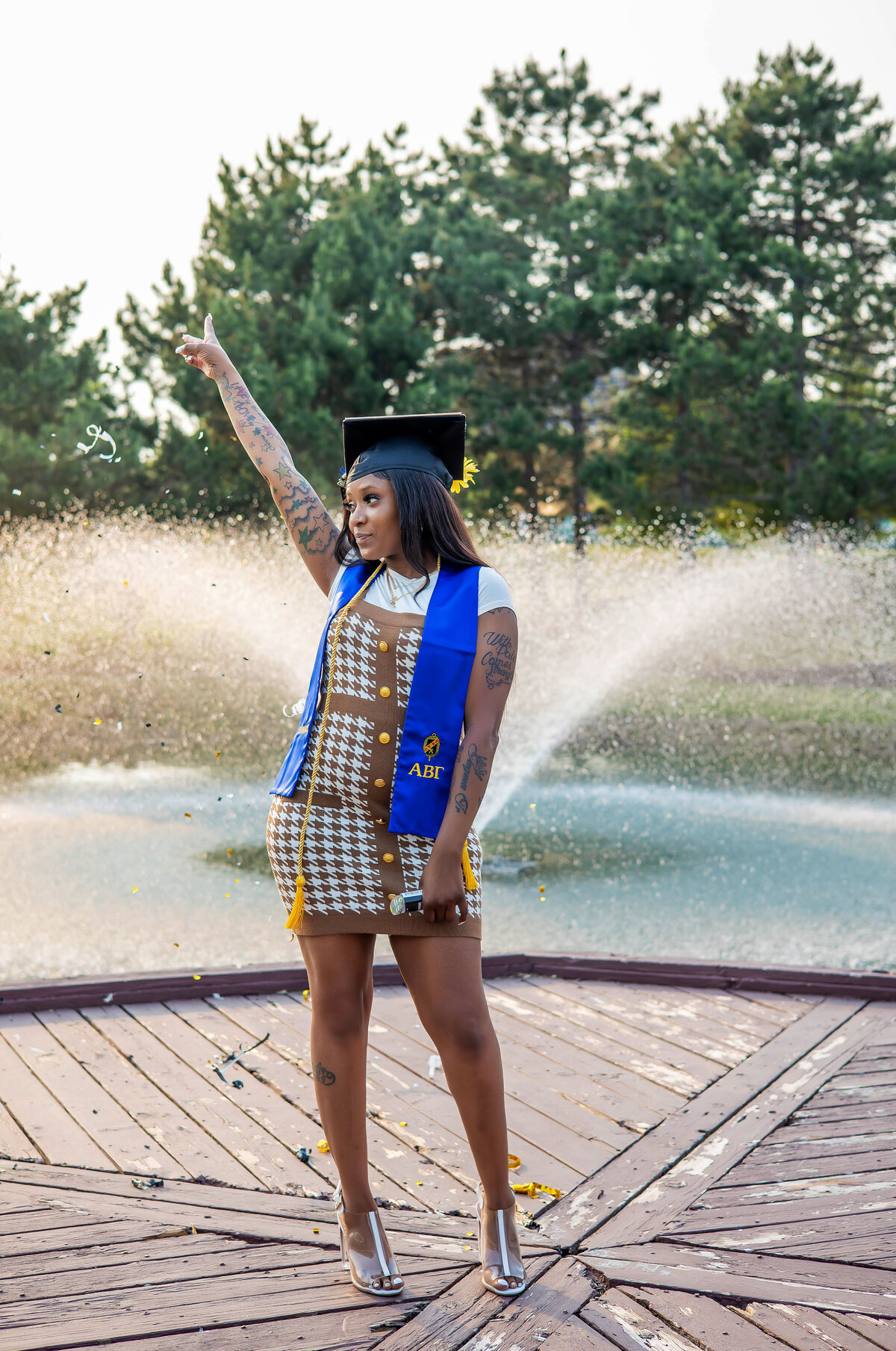 College graduate wearing black graduation cap and brown and white dress holding up the peace sign in front of a pond at sunset