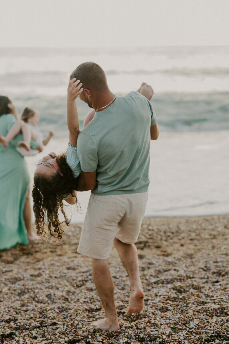 Papa habillé en vert et blanc faisant basculer sa grande fille dans les bras dans un jeu à la plage pour une séance photo famille en Vendée.