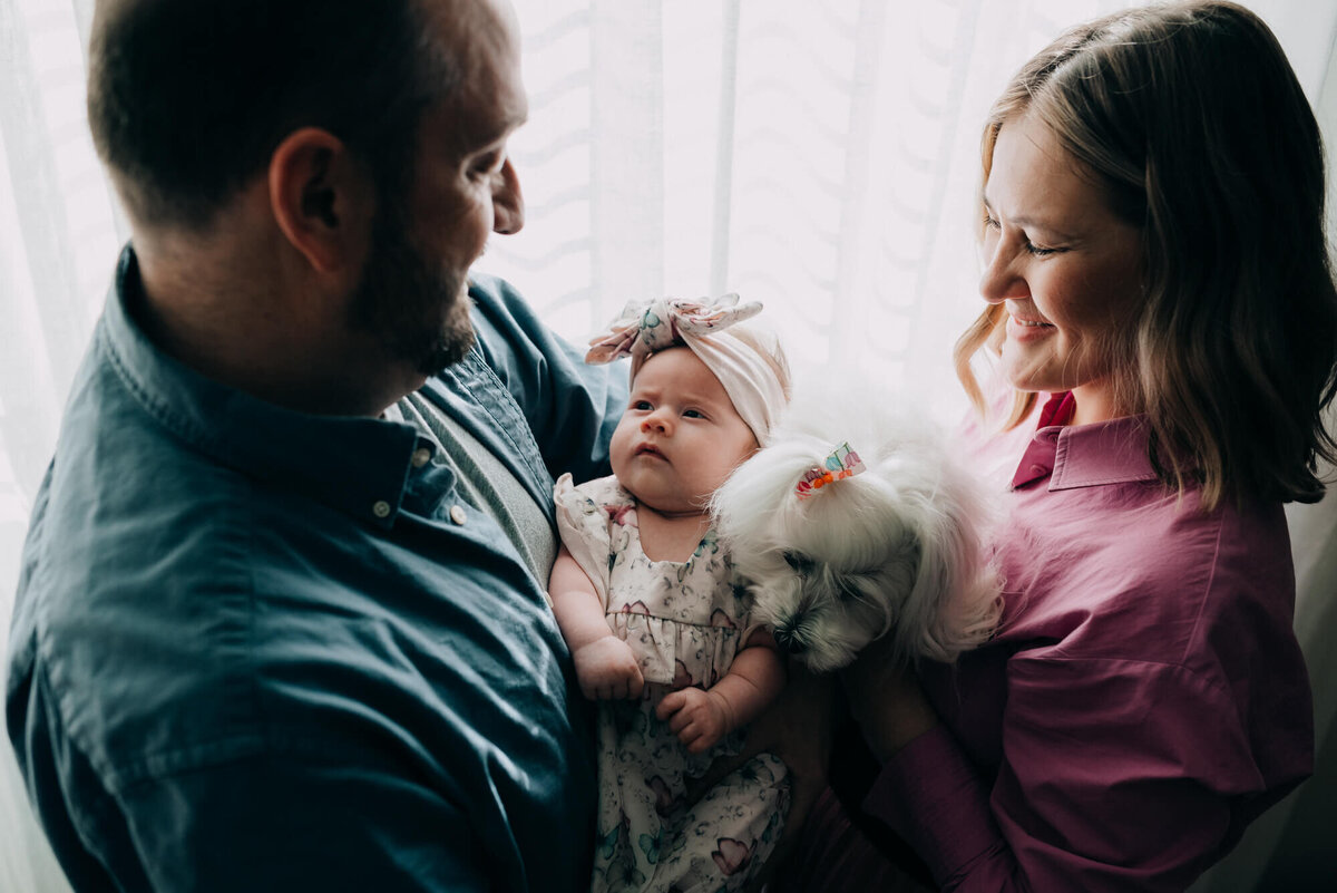 A mother and father hold their newborn daughter and their cute little white dog close together in front of a bright window