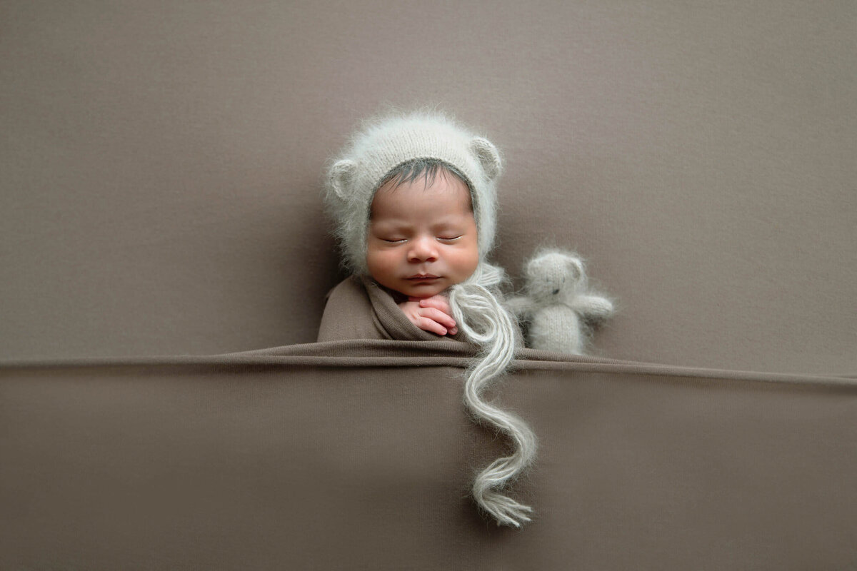 newborn baby boy wearing a brown hat holding a brown bear on a brown background.