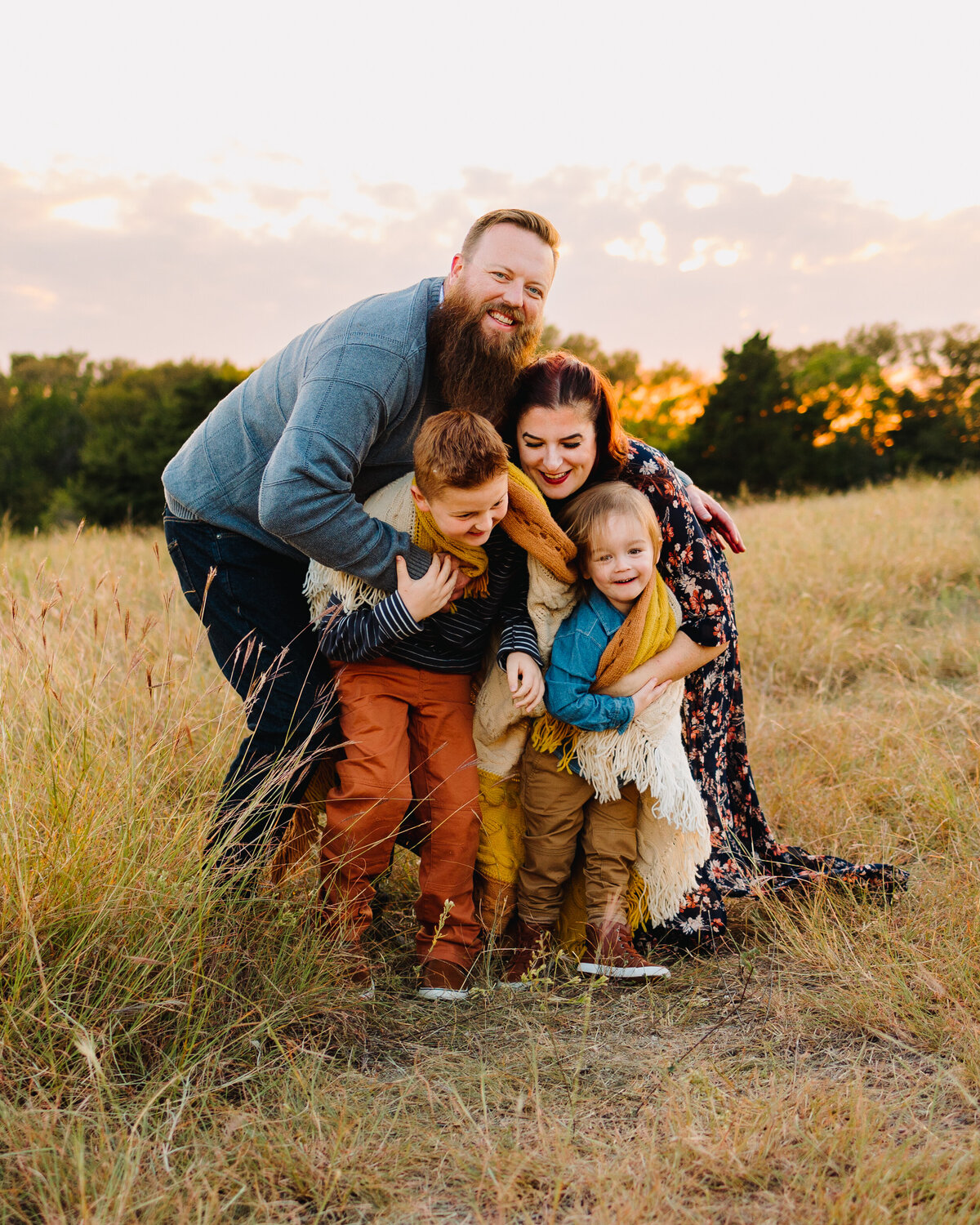 A family poses for a professional mini session in the fall. They are looking at the sunset and behind them are some trees. The dad is wearing a blue outfit and the mom is wearing a dress with flowers, and the kids have a scarf around their necks.