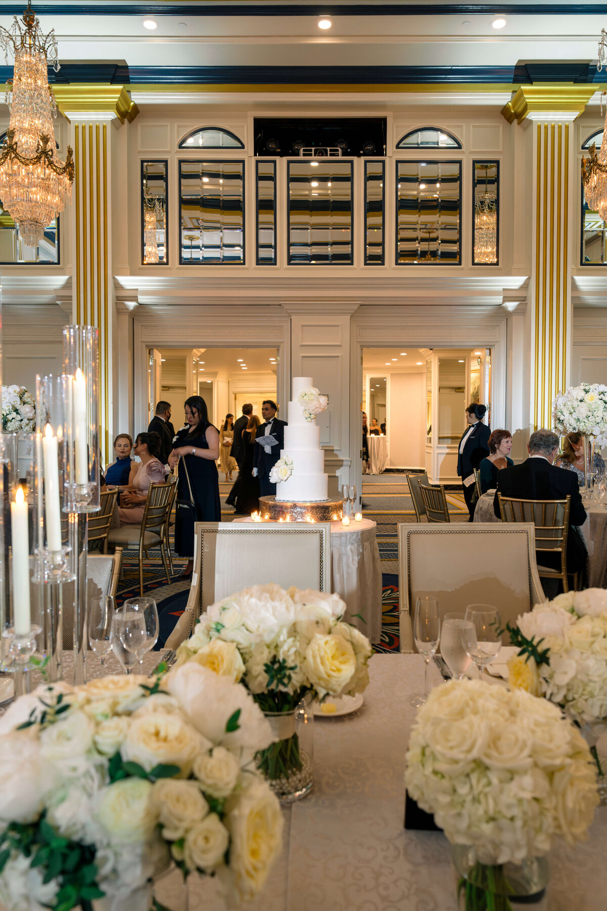 Elegant wedding reception with a large tiered white cake. Guests in formal attire are seated at tables adorned with white floral arrangements and tall candles. The room features chandeliers, gold accents, and mirrored walls.