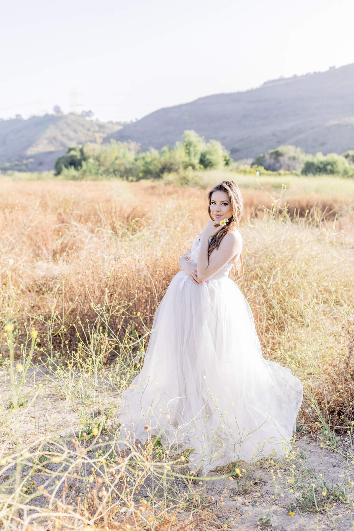 A woman in a flowing white gown stands in a sunlit field of tall grasses, with rolling hills in the background. She holds a bouquet of yellow flowers and smiles at the camera, creating a serene and whimsical scene.