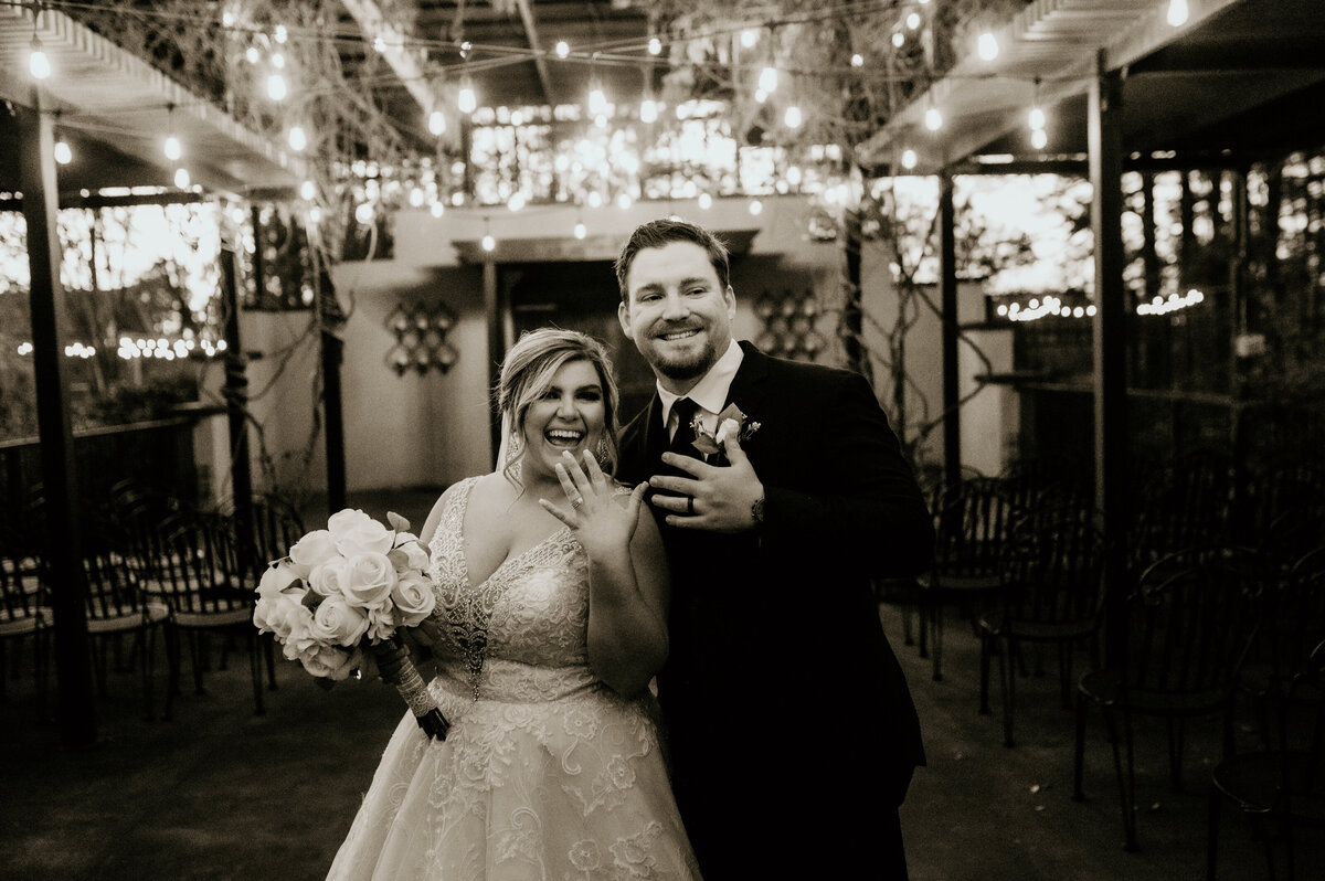 bride and groom holding up their hands showing off their wedding rings and smiling under the string lights