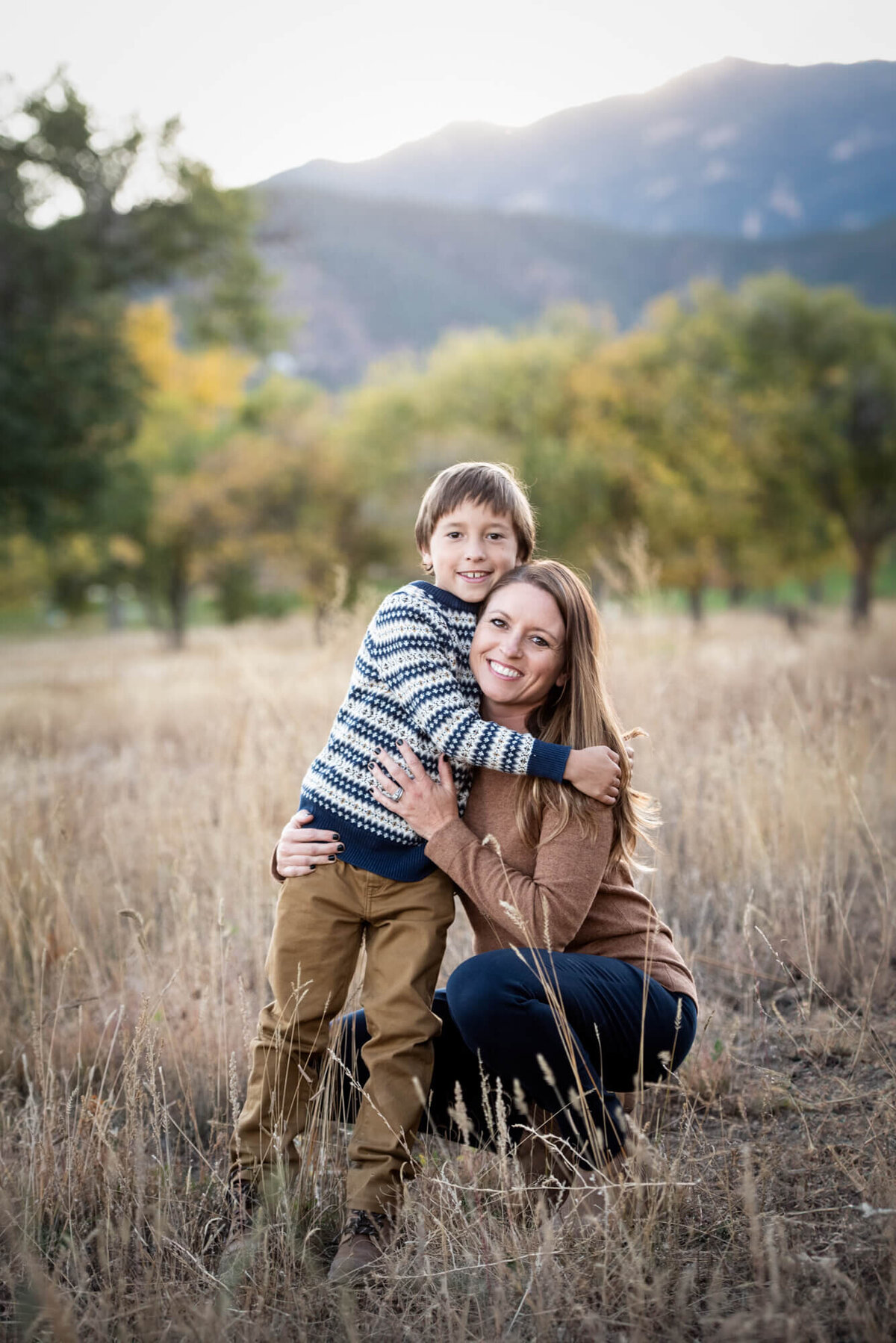A mother in a brown shirt hugs her young son in a field at sunset
