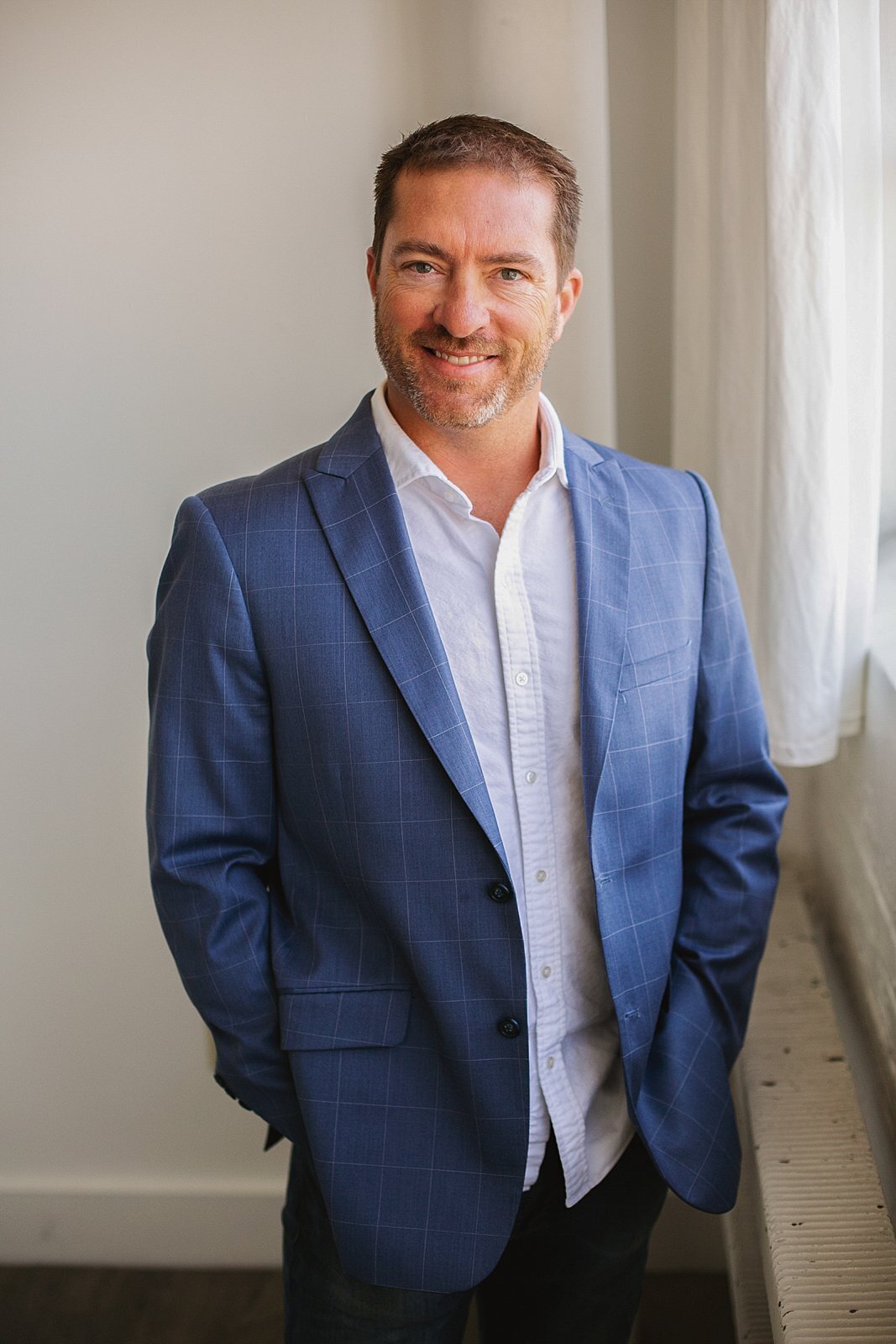 a-smiling-business-professional-in-a-blue-checkered-blazer-standing-in-front-of-a-bright-window-for-a-linkedin-headshot-in-indianapolis