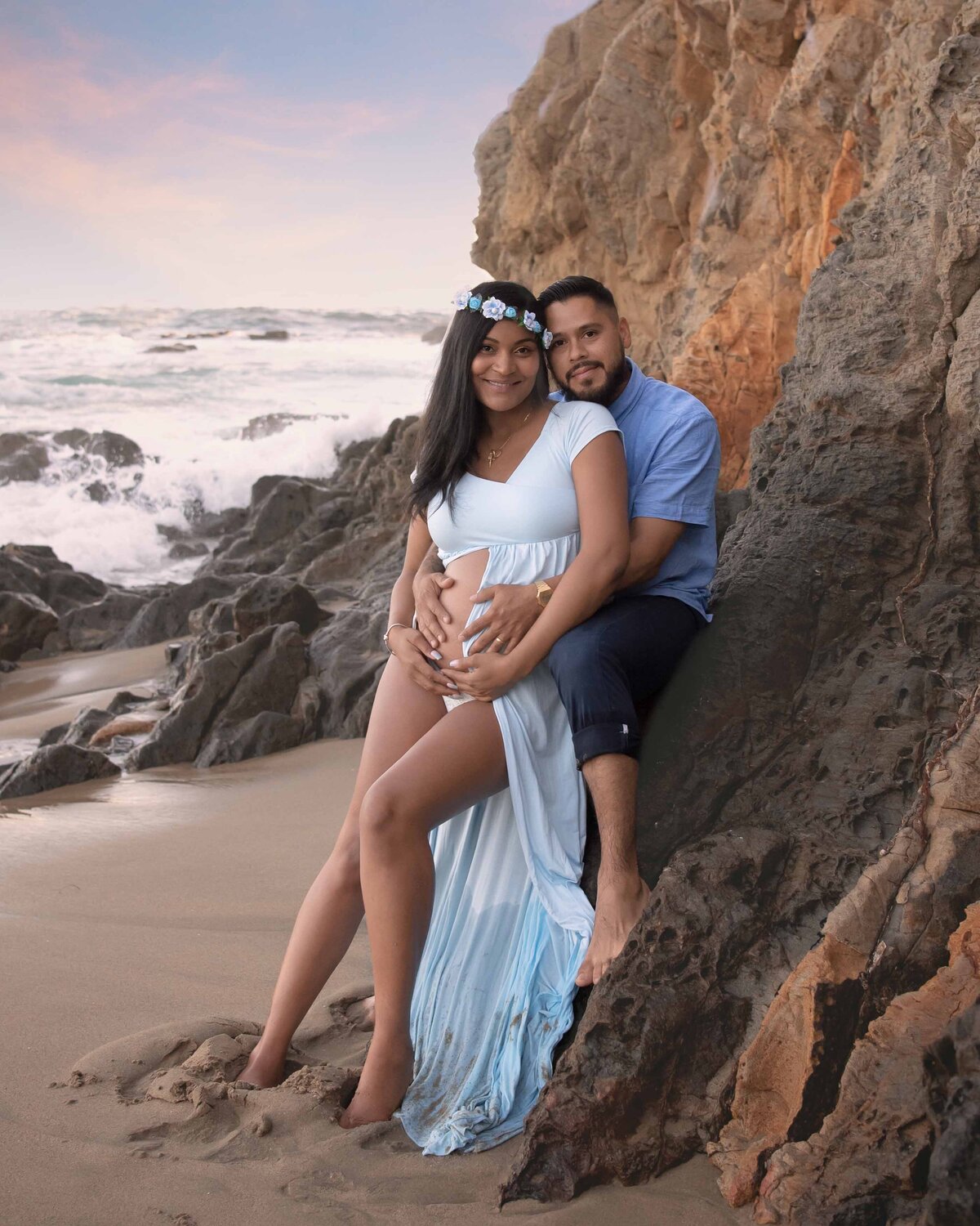 Expectant Mom and Dad on California beach sitting on rocks holding baby bump.