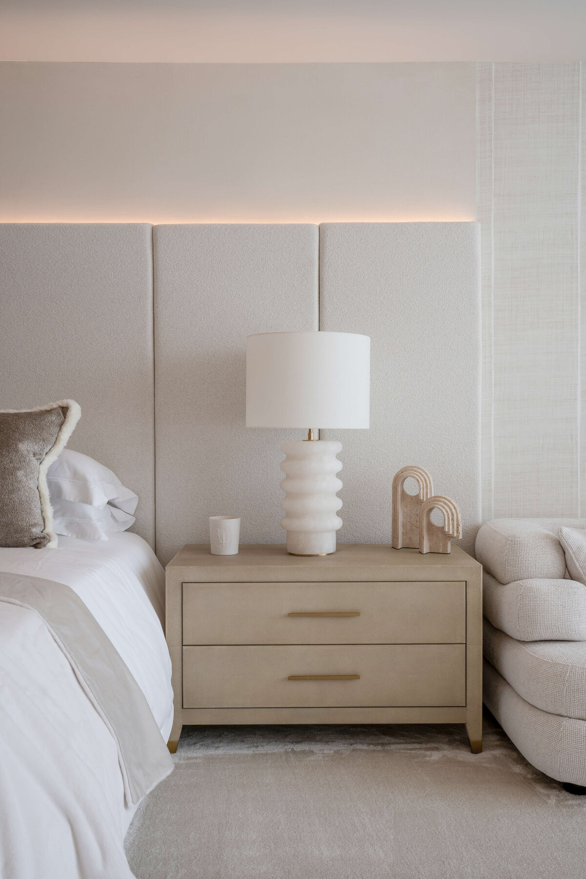 A light, neutral bedside table between the bed and couch in the primary bedroom. Atop the table are a chic white lamp and other neutral accessories.