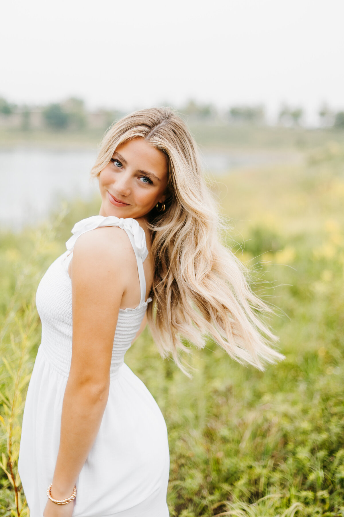 Senior girl in a white dress twirling with her hair blowing behind her in front of a grassy field