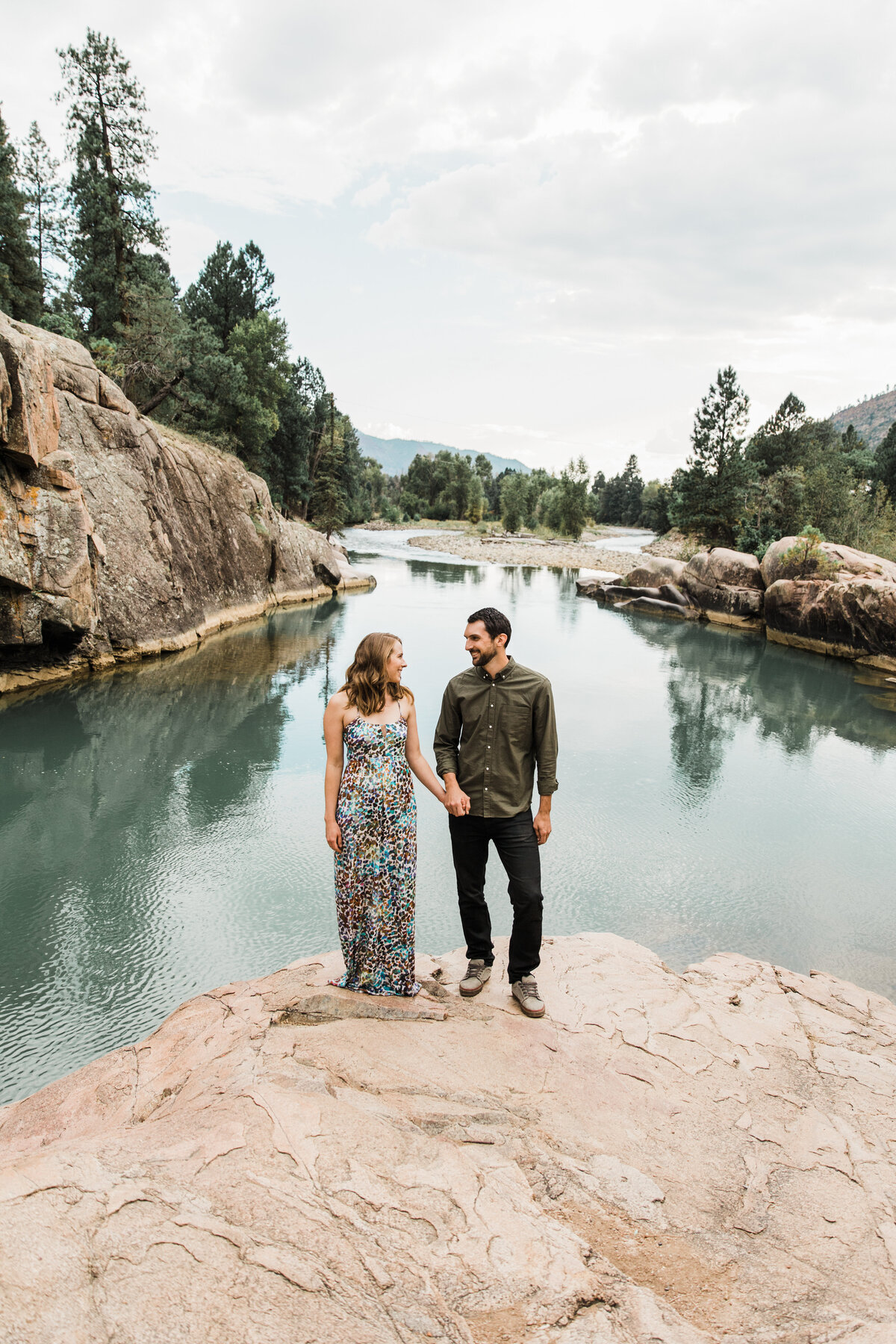 A couple holding hands and standing on a large rock in front of the Animas River during their engagement session in Durango, Colorado. The woman on the left is wearing a long, vibrant, colorful dress. The man on the right is wearing a dark green dress shirt and dark pants. Trees. rocks, and mountains can be seen behind them beyond the river.