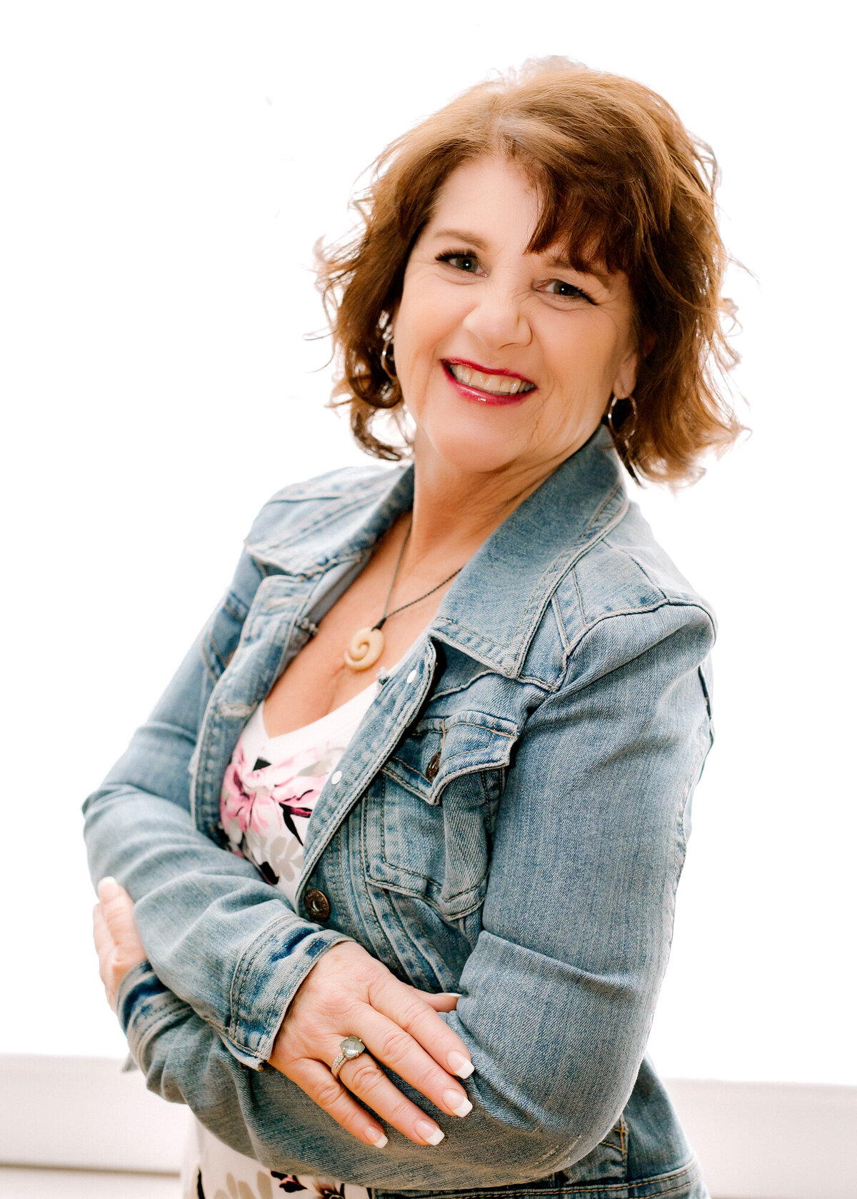 Mature female headshot client wearing a jean jacket & floral top with arms crossed, smiling at the camera