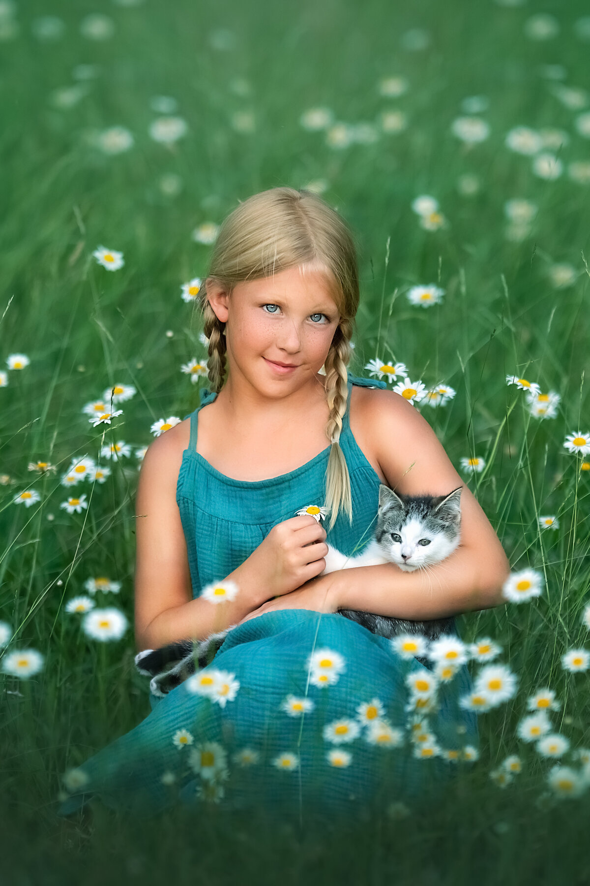 blue eye blond girl in teal dress holding a kitten and sitting in the field of daisy flowers.