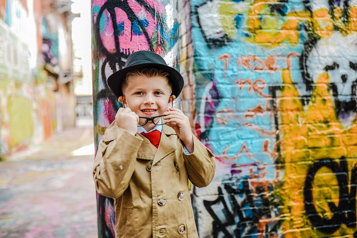 Little boy in a black fedora and trench coat taking of glasses in graffiti alley near MICA in Baltimore Maryland