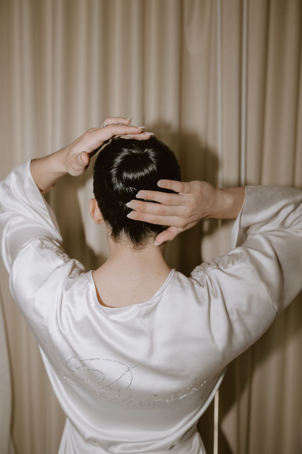 bride reaching towards slick back bun in front of tan curtain wearing ivory silk robe