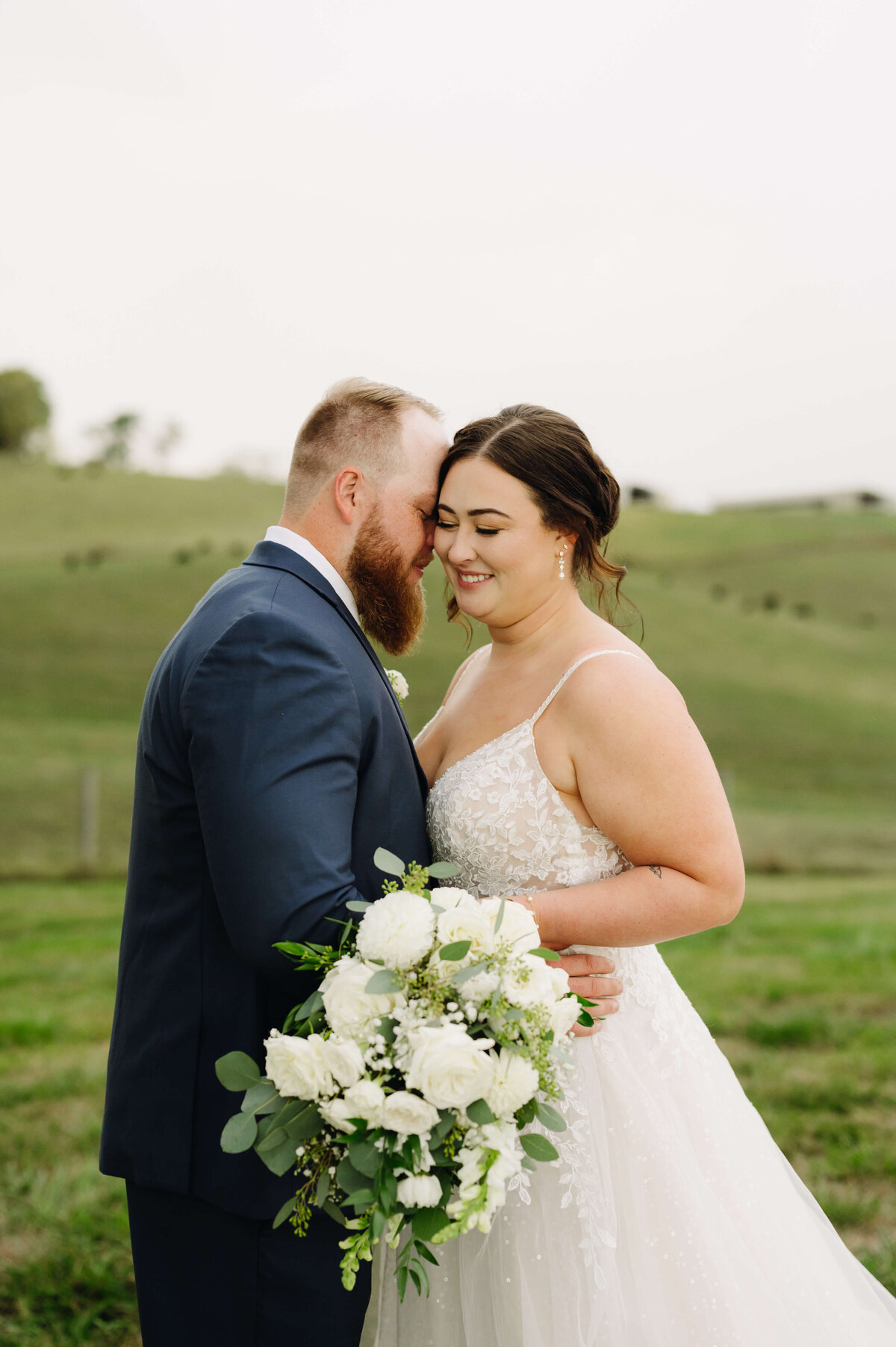 wedding portrait of groom leaning into his bride and whispers to her as she smiles while holding a white rose bouquet at Sunny Slope Farm