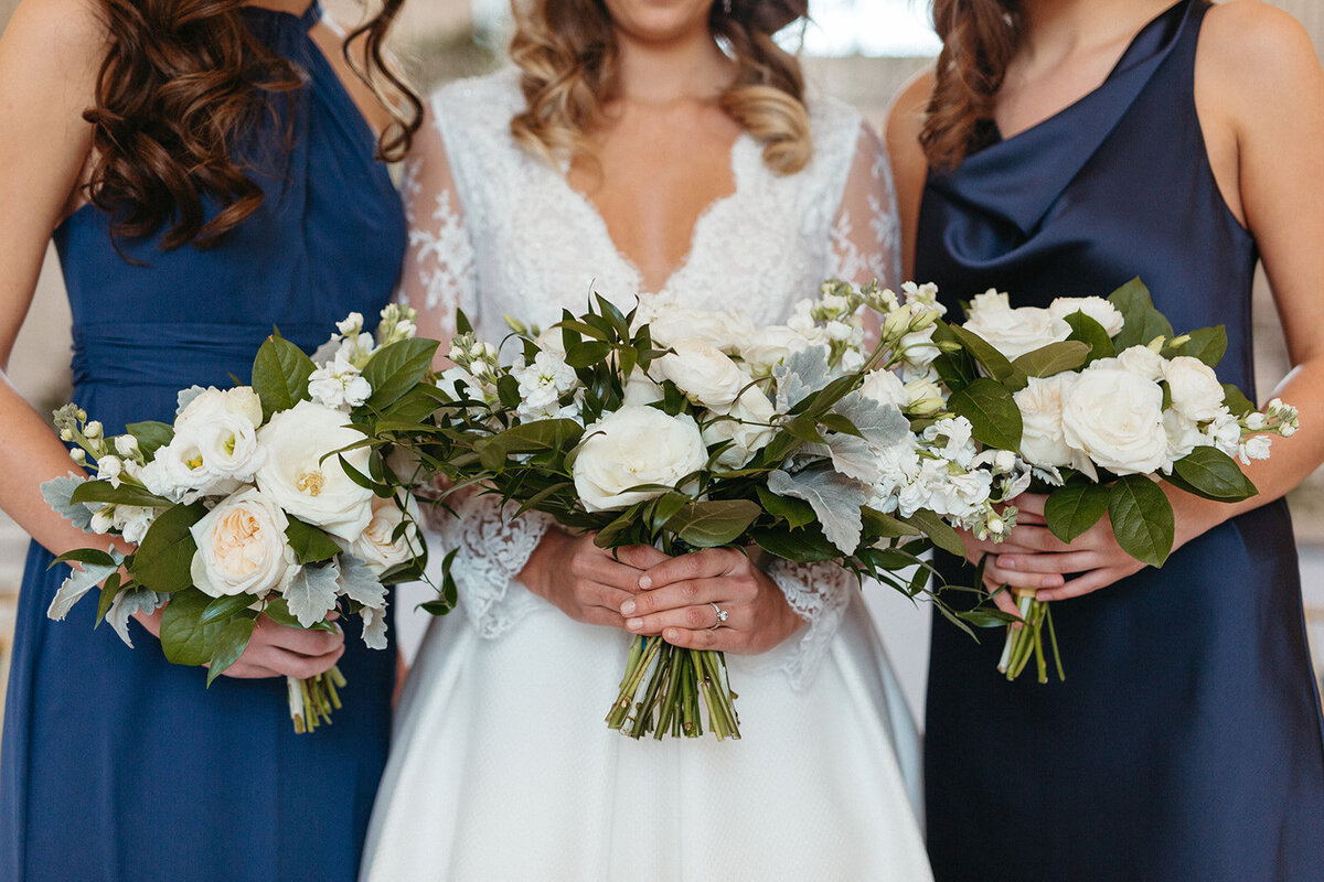 A bride in a white wedding gown and two bridesmaids in blue gowns holding a white bouquet of flowers.