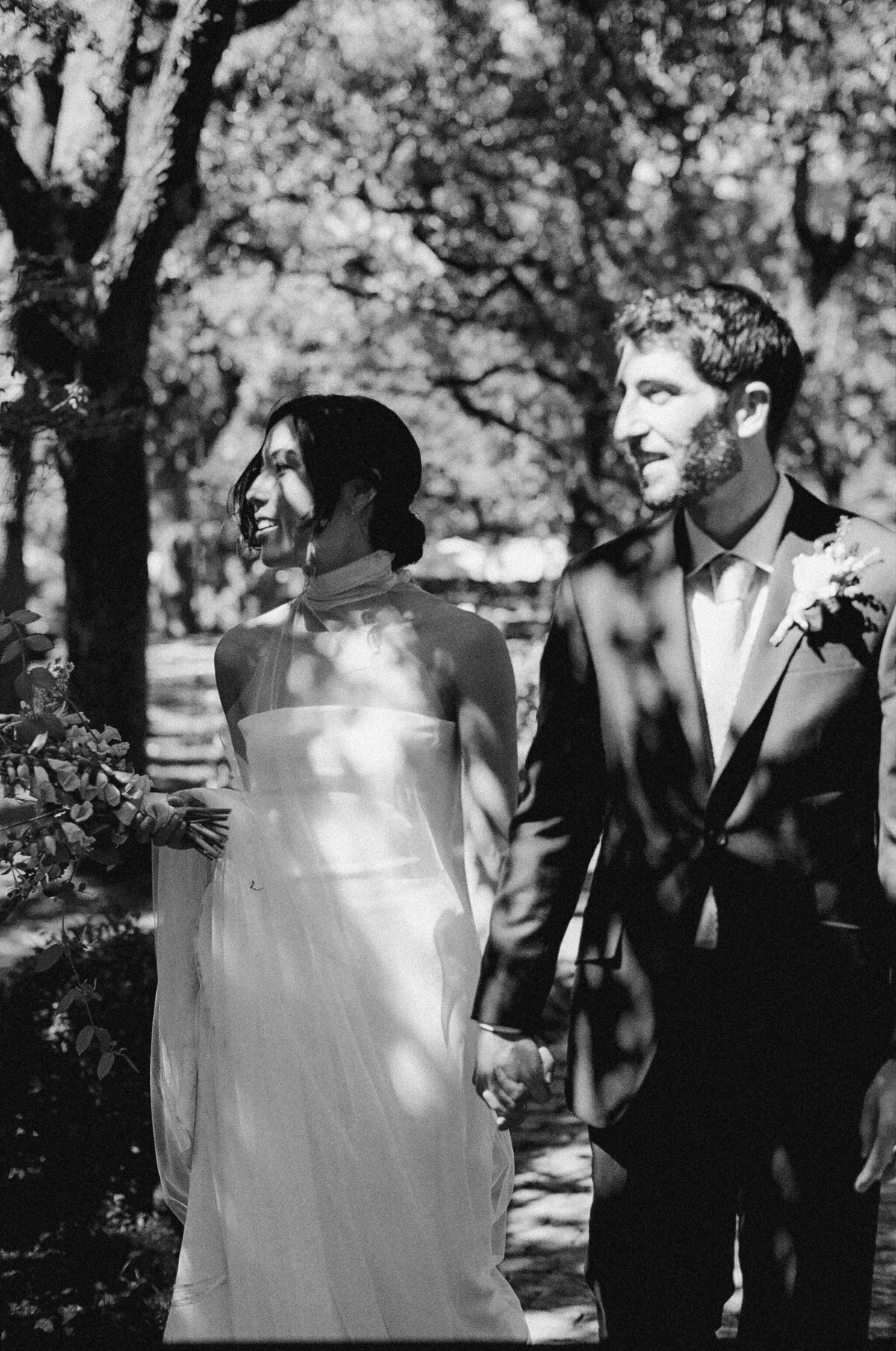 Black and white image of bride and groom walking through trees at Matties wedding venue Austin