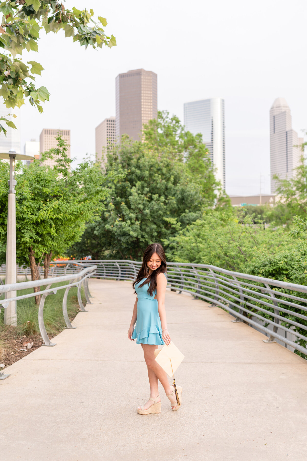 Houston high school senior girl wearing baby blue dress and holding cap down at side and looking at it with Downtown Houston in the background